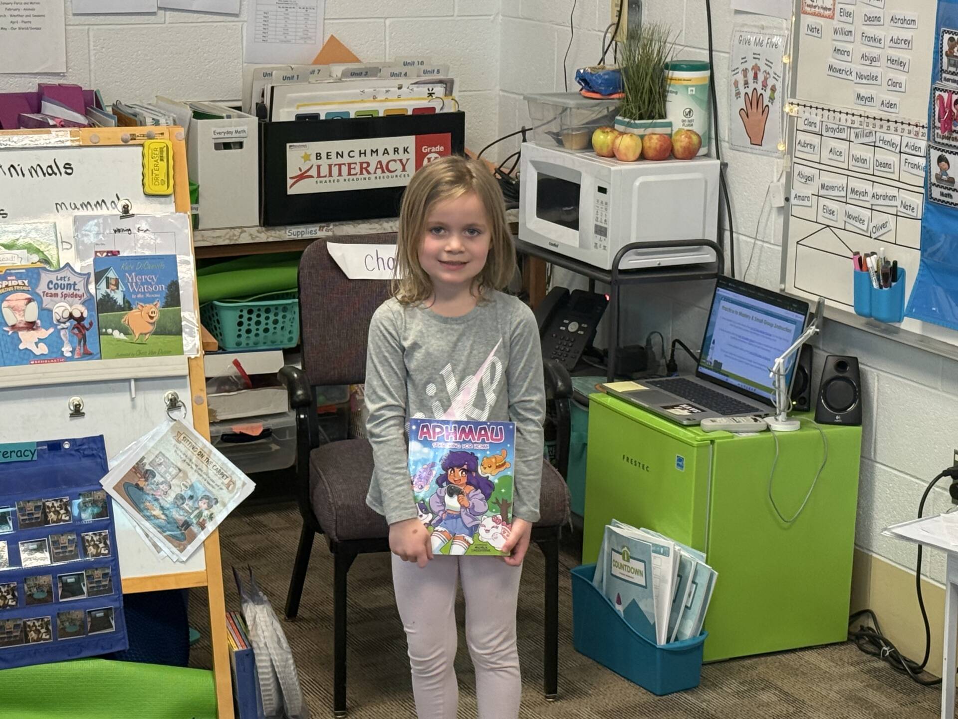 Young student stands in a classroom holding an “Aphmau” book, surrounded by reading materials and classroom supplies.