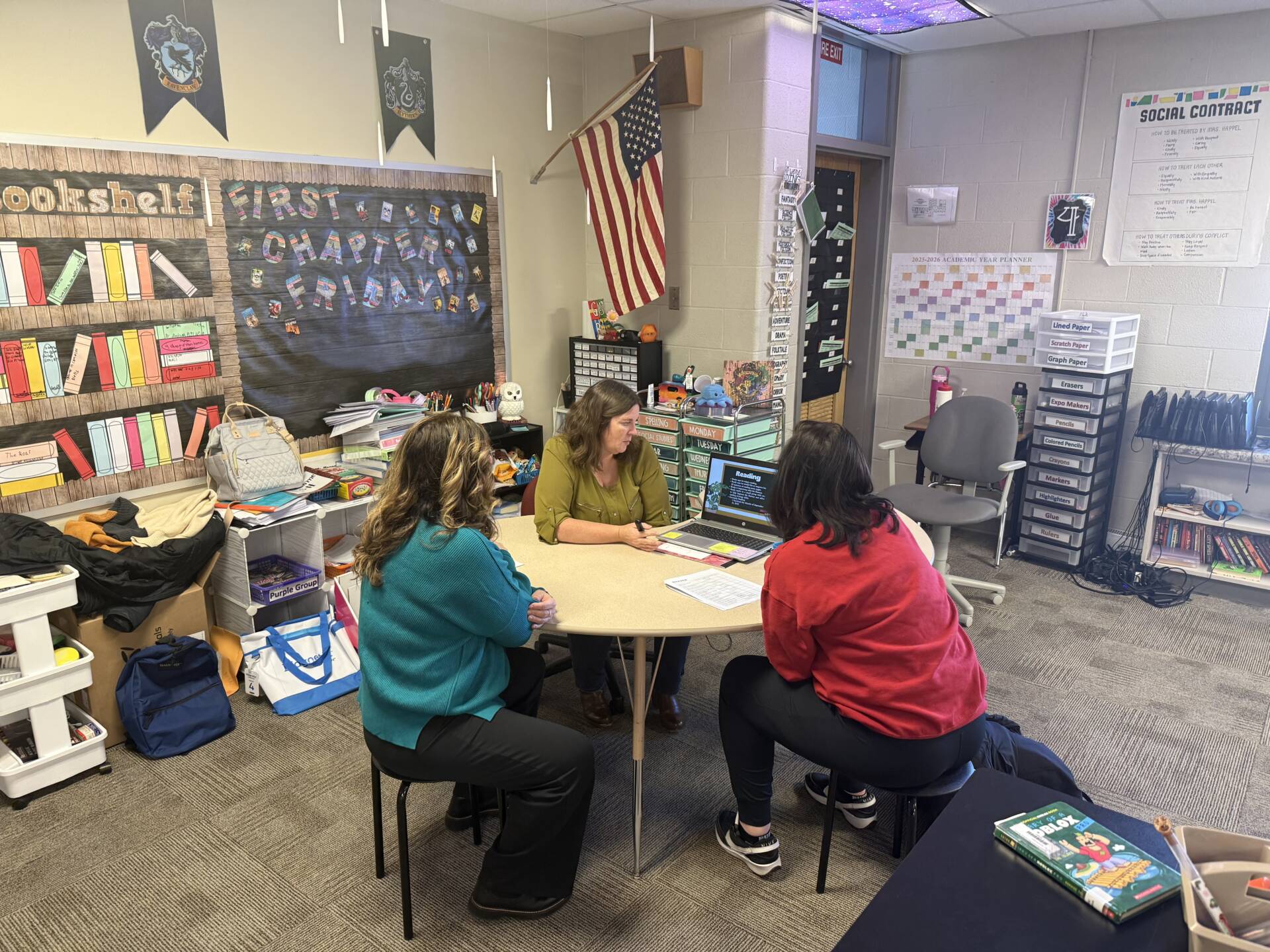 Three adults sit around a table in a classroom reviewing materials on a laptop during a small meeting.