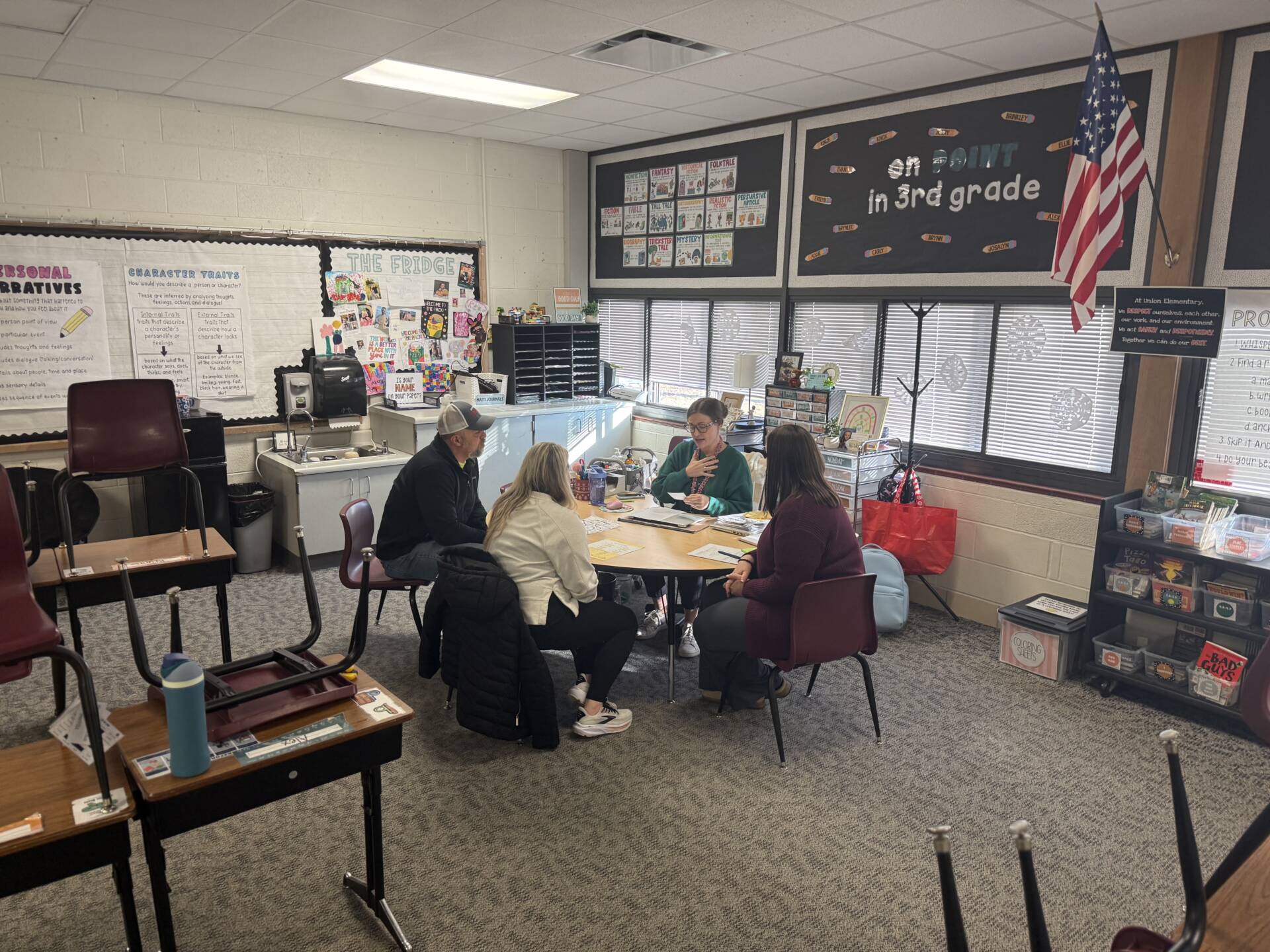 Teacher meets with three adults around a table in a third-grade classroom with posters, books, and an American flag nearby.