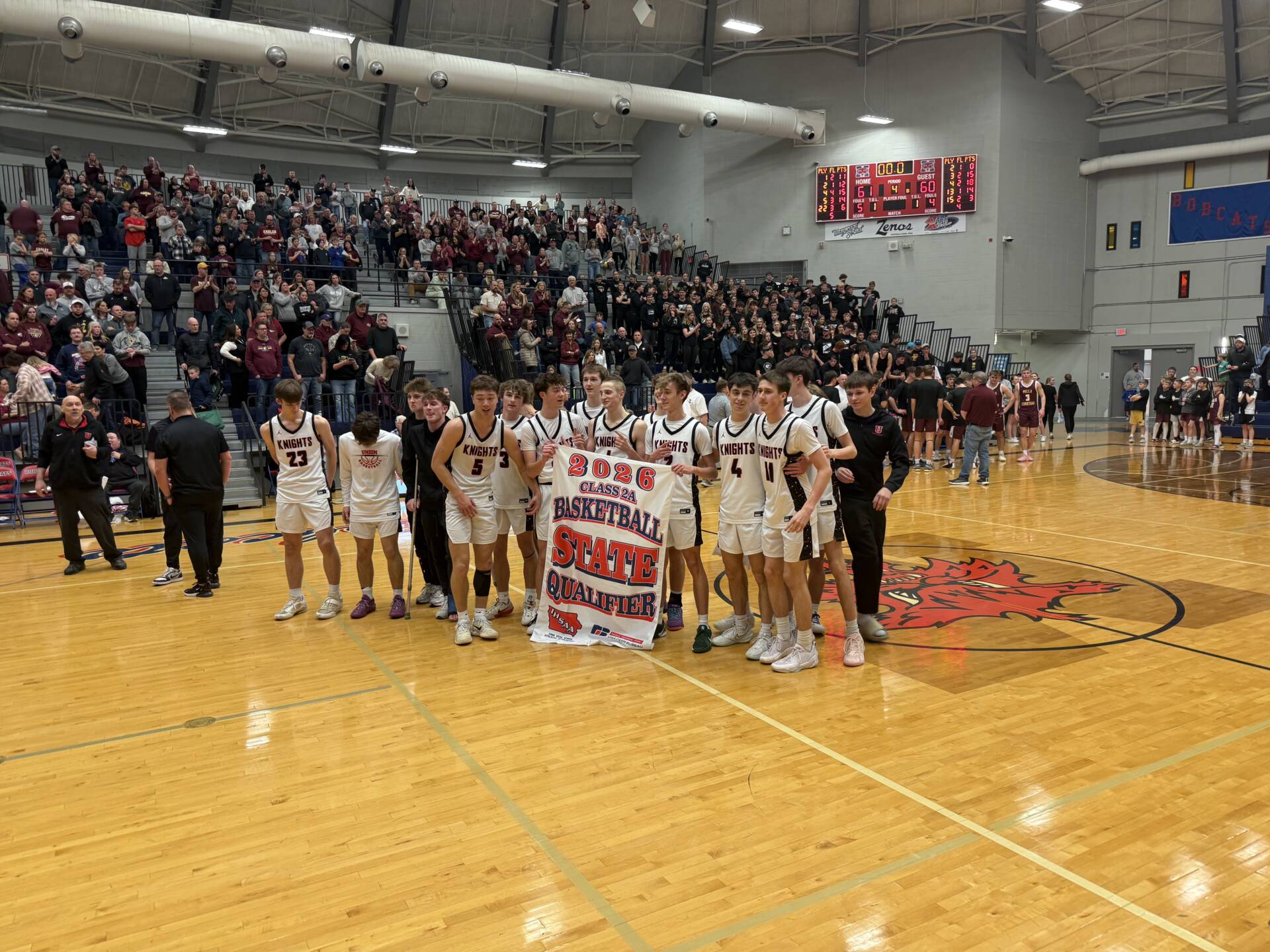 High school basketball team stands on the court holding a “2026 Class 2A Basketball State Qualifier” banner as a crowd cheers in the gym.