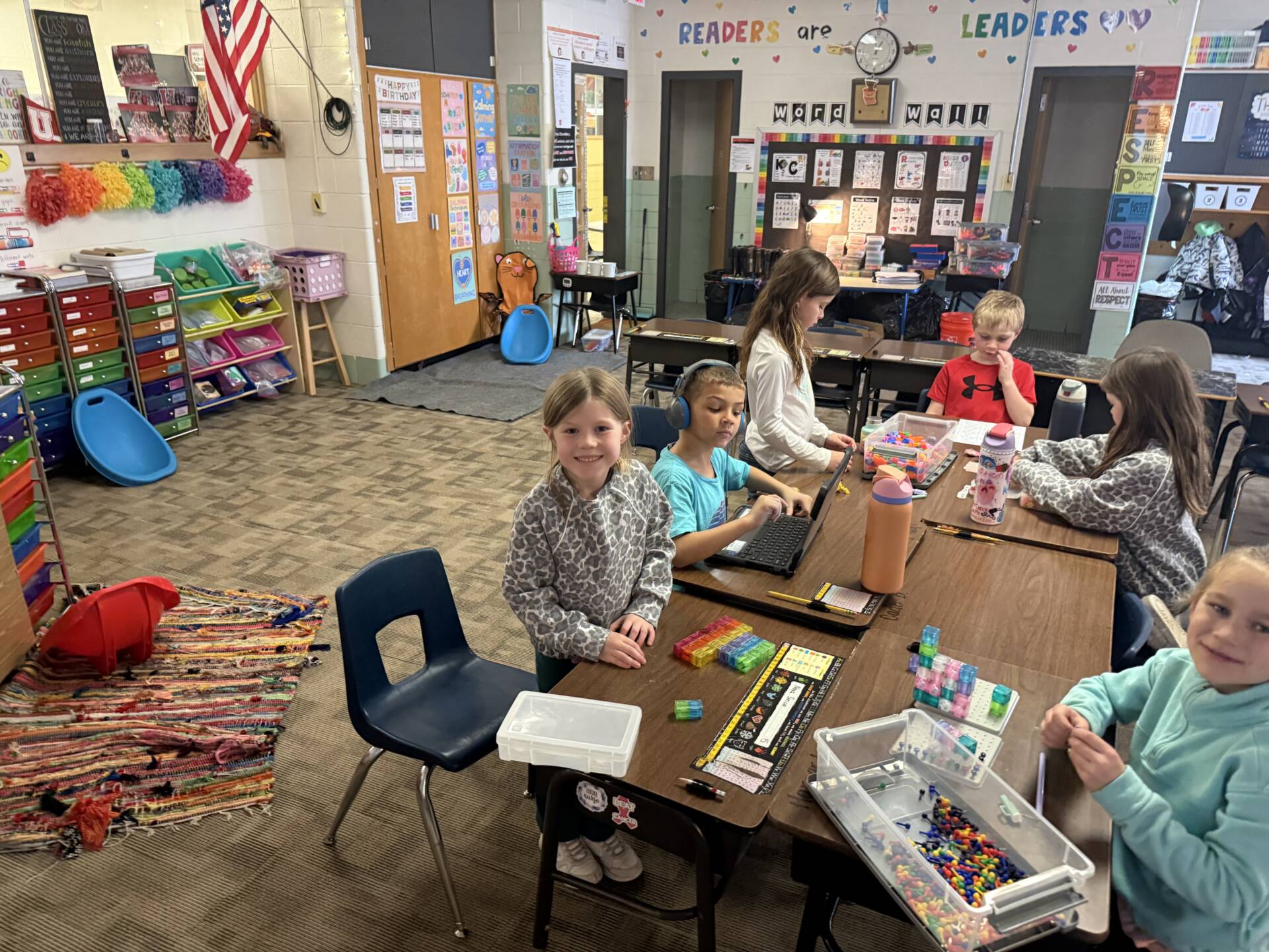 Elementary students sit at desks using small building blocks and a laptop during a classroom learning activity.