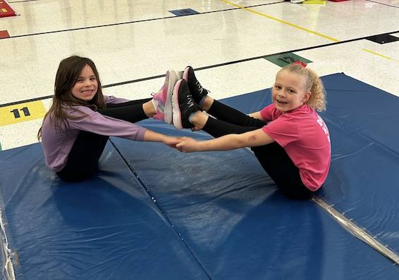 Two elementary students sit on a gym mat balancing with their feet together while holding hands during a PE activity.