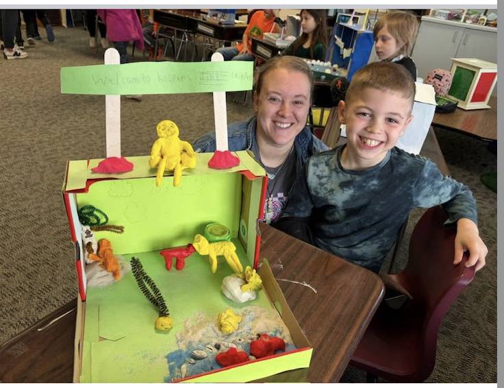 Student and adult smile beside a colorful diorama project displayed on a classroom desk.