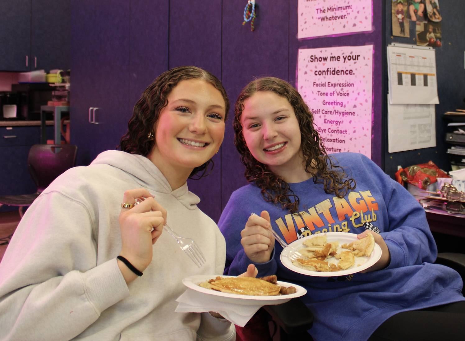 Two high school students smile while holding plates of pancakes during a classroom breakfast activity.