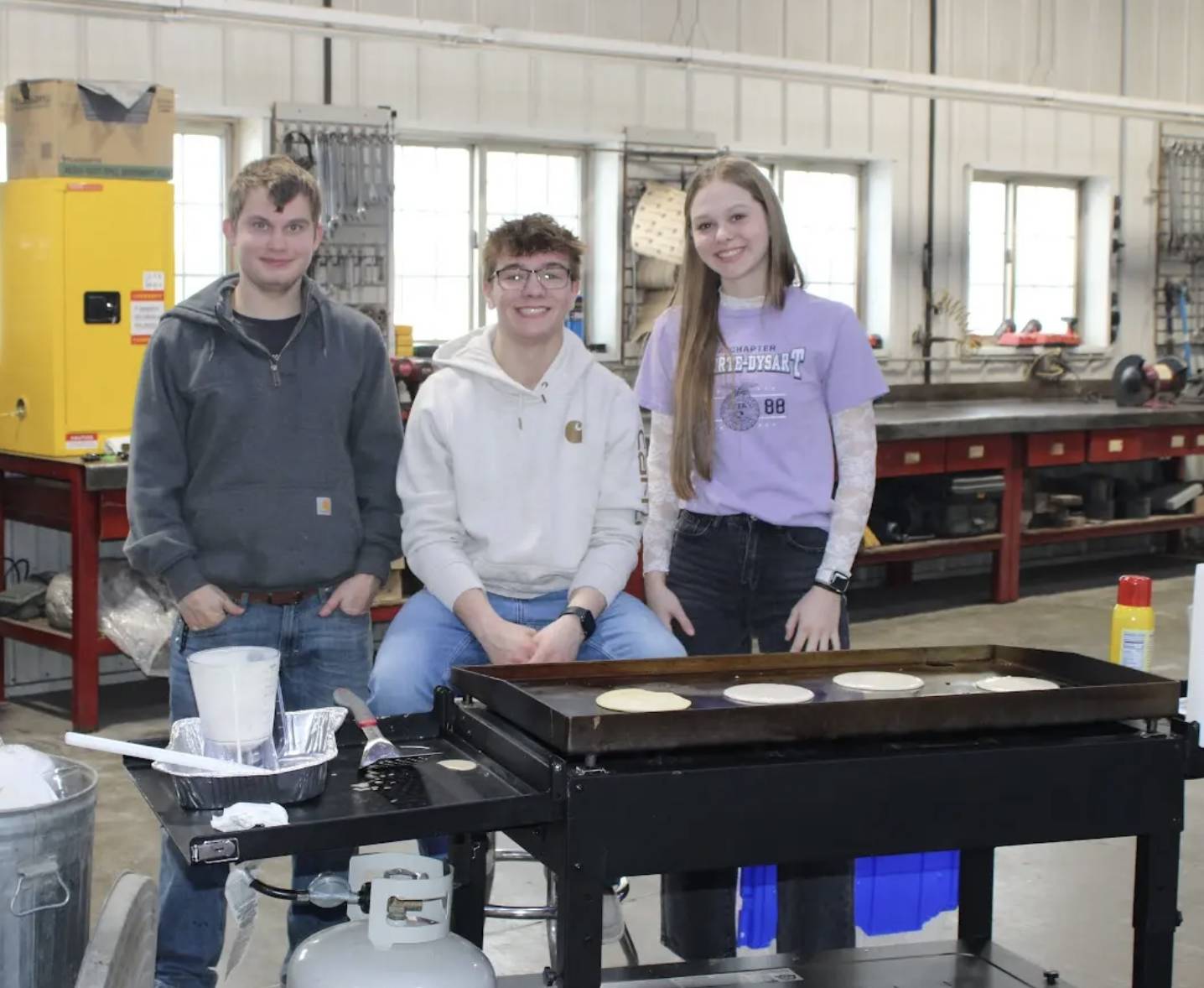 Three high school students stand beside a griddle cooking pancakes in an agricultural or shop classroom.