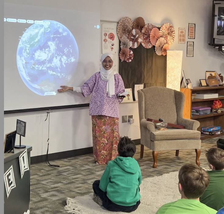 A guest presenter stands beside a projected image of Earth while speaking to students seated on the floor.