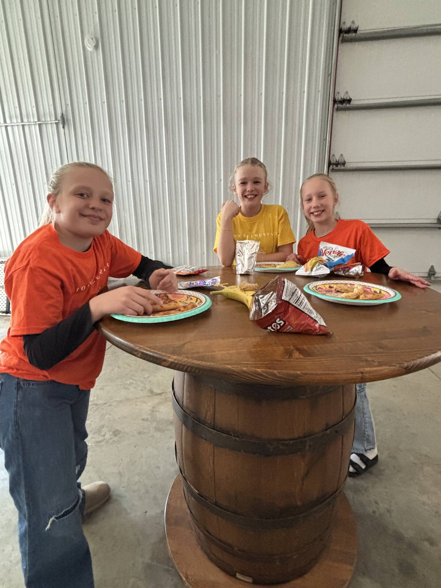 Three girls sit at a round wooden table eating snacks and smiling inside a barn-style room.