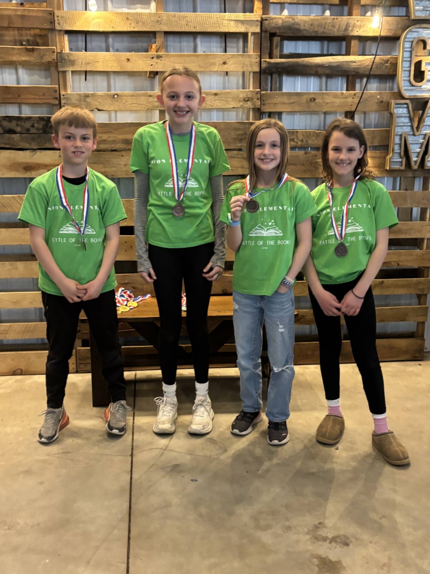 Four students wearing green shirts stand with medals, smiling after a “Battle of the Books” event.