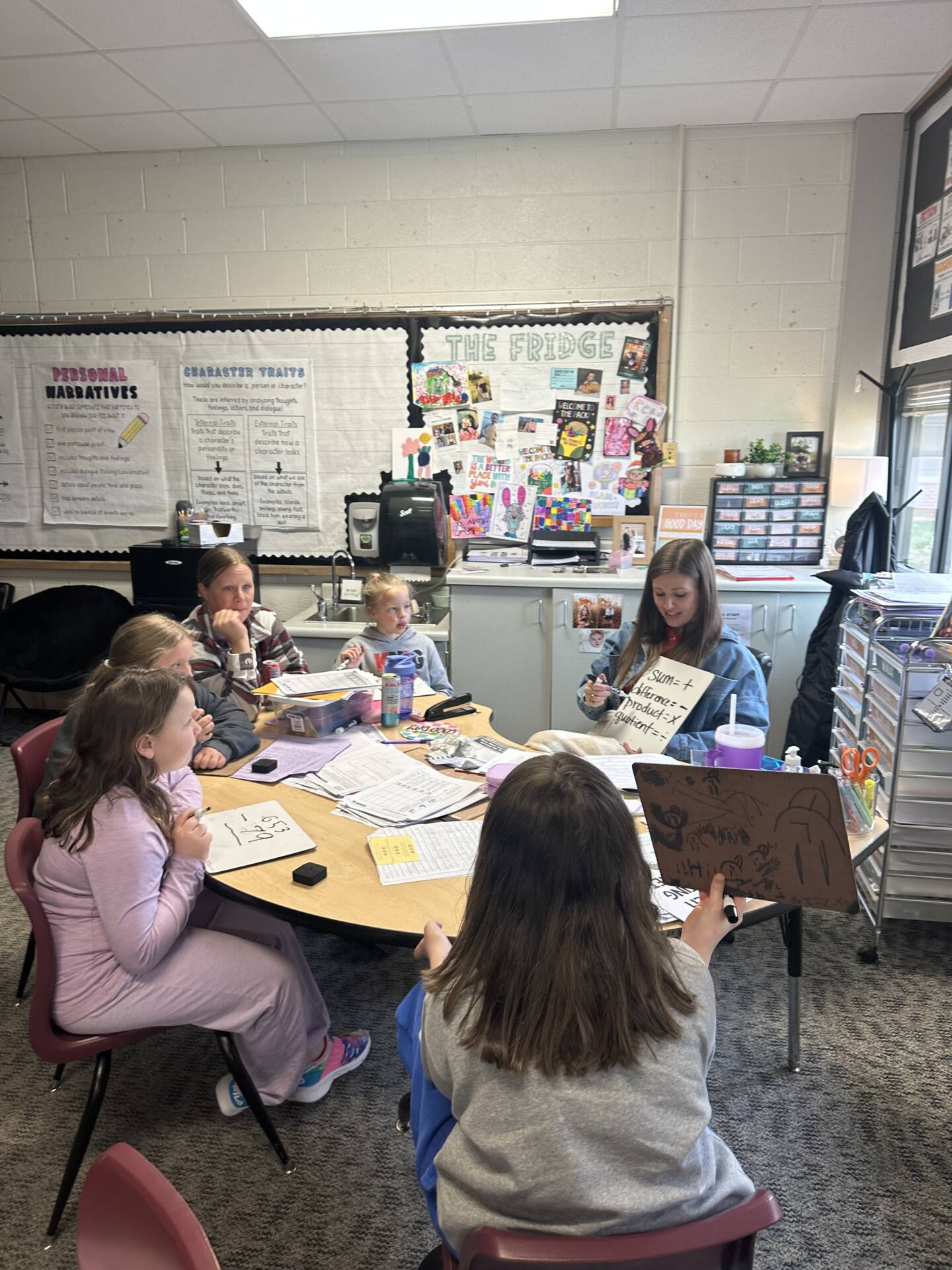 Teacher leads a small math group at a round table as students use whiteboards and worksheets in a classroom.