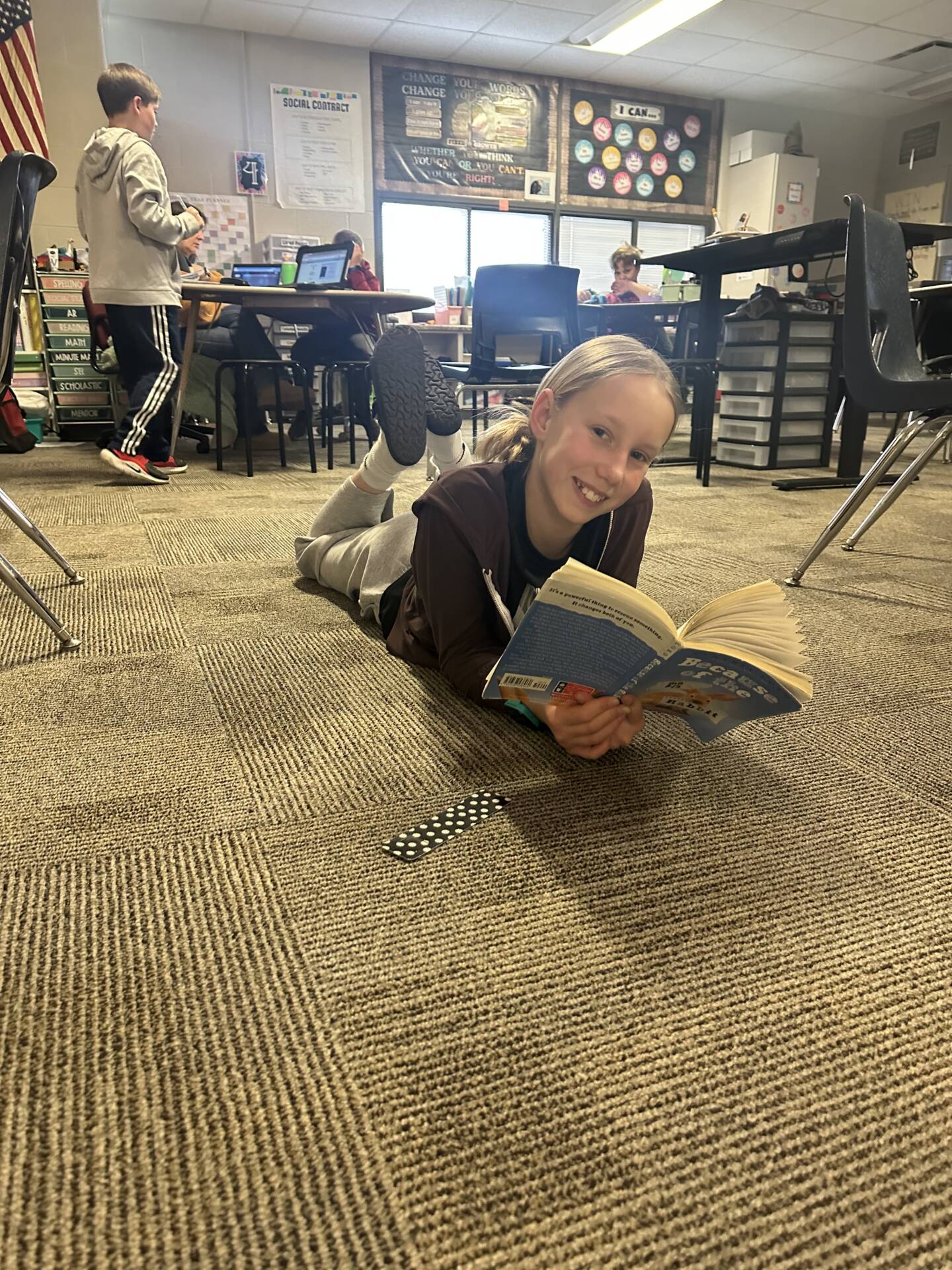 Student lies on classroom carpet reading a book, smiling at the camera while classmates work at desks behind.