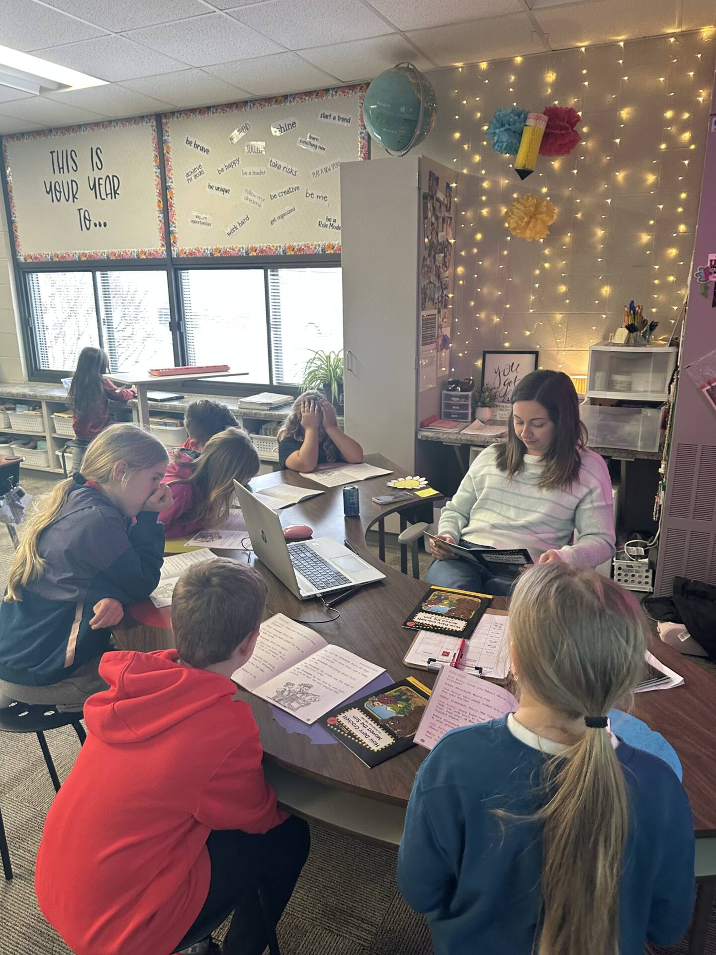 Teacher reads to a small group of students gathered around a table in a cozy classroom with lights and posters.