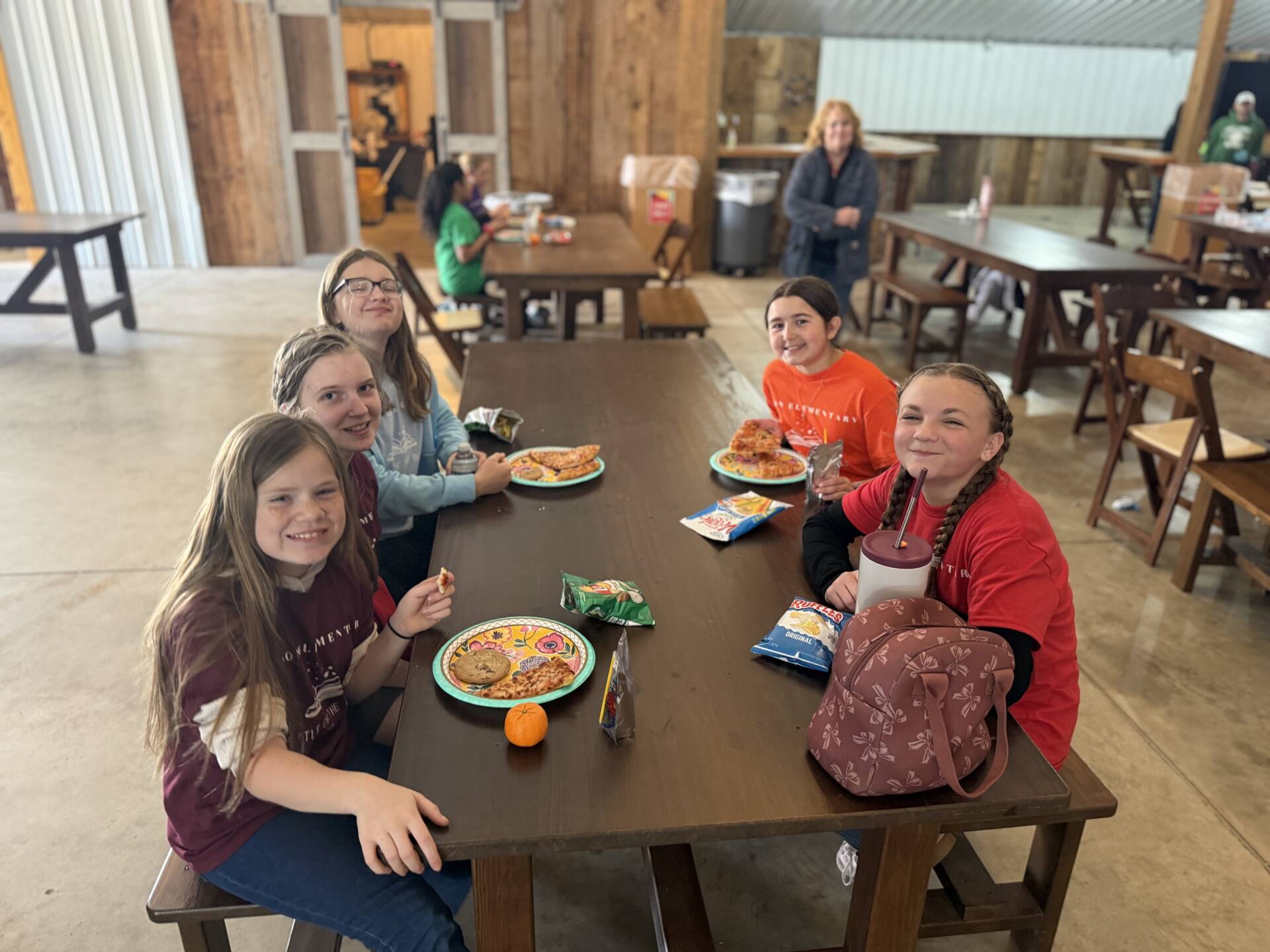 Five girls sit at a long wooden table in a barn-like space, smiling while eating pizza, chips, and snacks