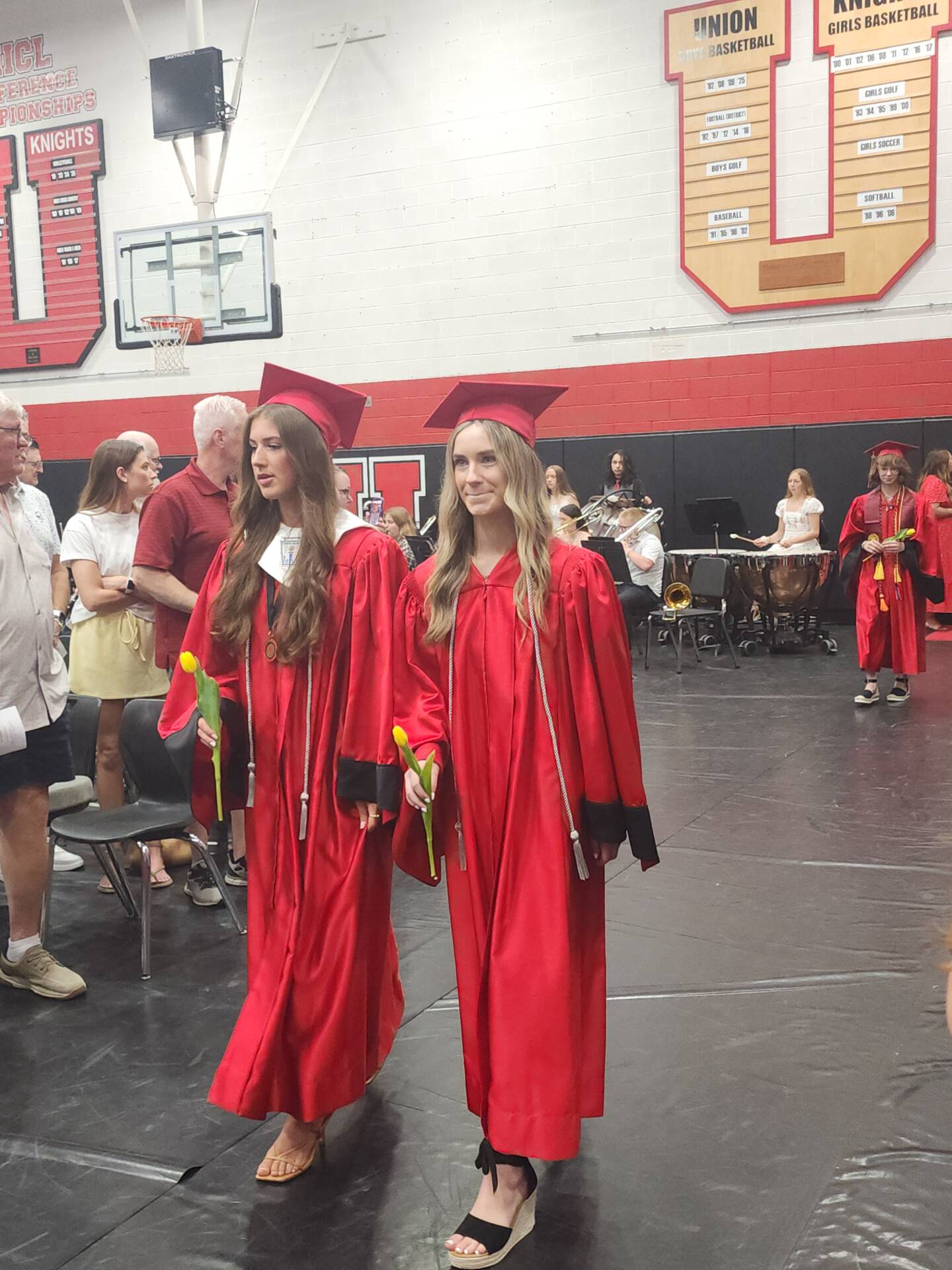 Two graduates in red caps and gowns carry yellow tulips while entering the graduation ceremony.
