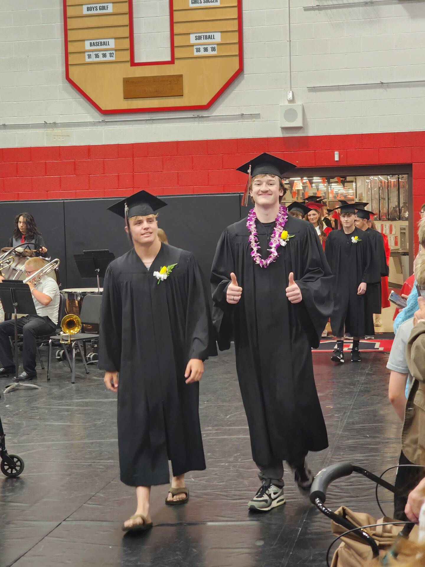 Two graduates in black caps and gowns walk into commencement, one wearing a purple lei and giving thumbs up.