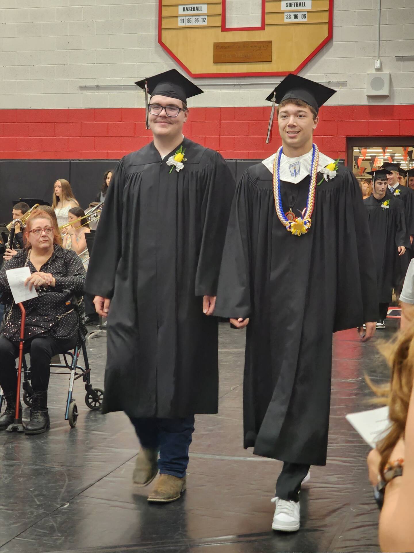 Two graduates in black caps and gowns walk into the gymnasium during the graduation ceremony.