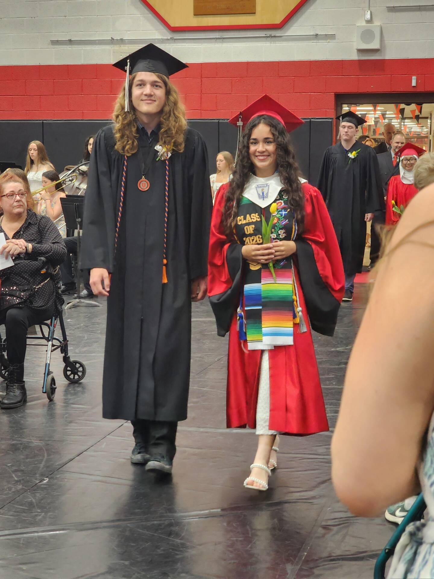 Graduate in a red cap and gown with a colorful stole walks beside a graduate in black during commencement.