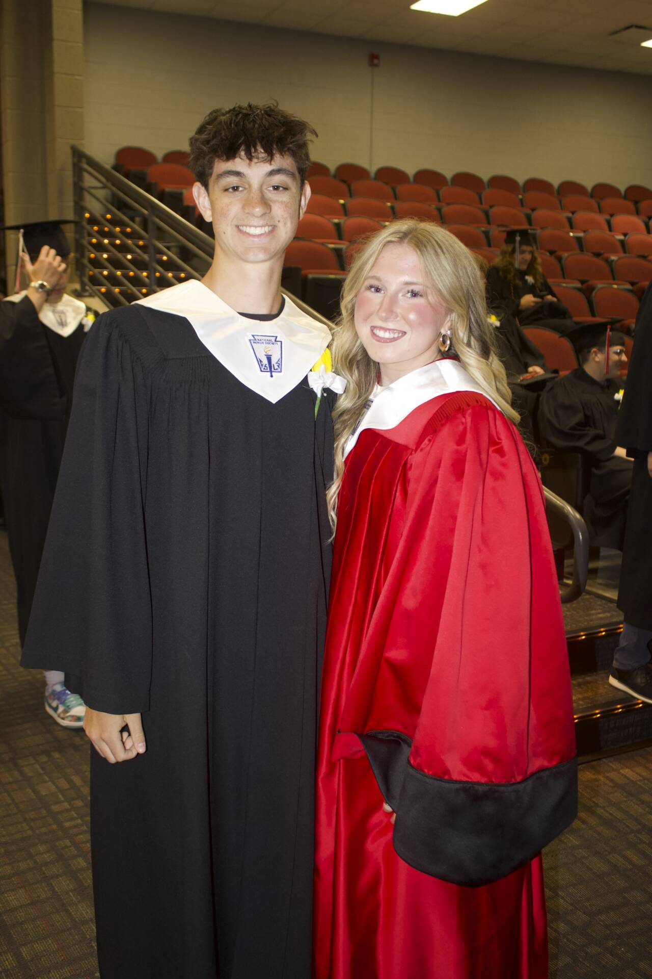 Graduate in a black gown and graduate in a red gown pose together inside the auditorium before commencement.