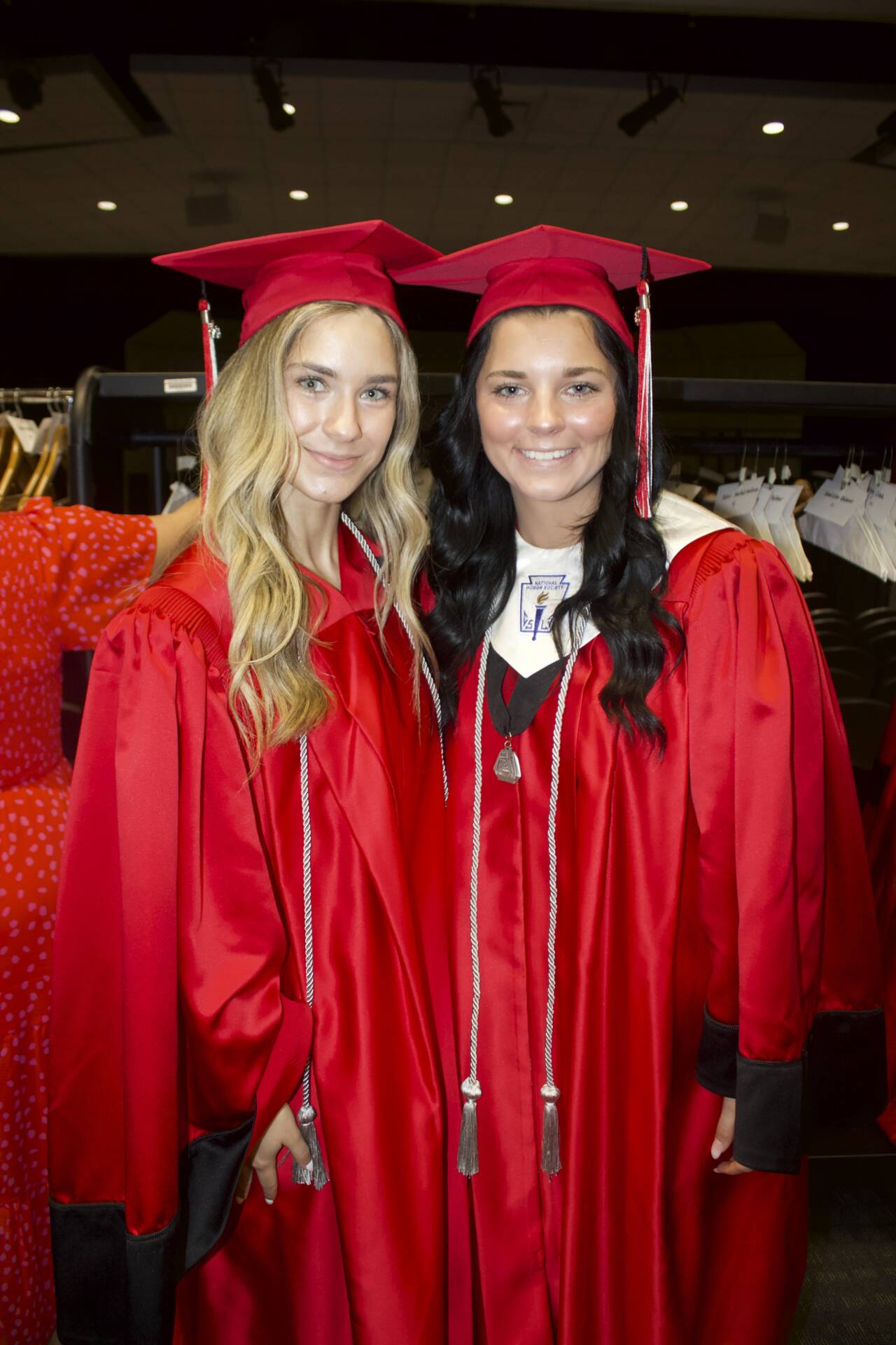 Two graduates in red caps and gowns smile for a photo before the graduation ceremony.