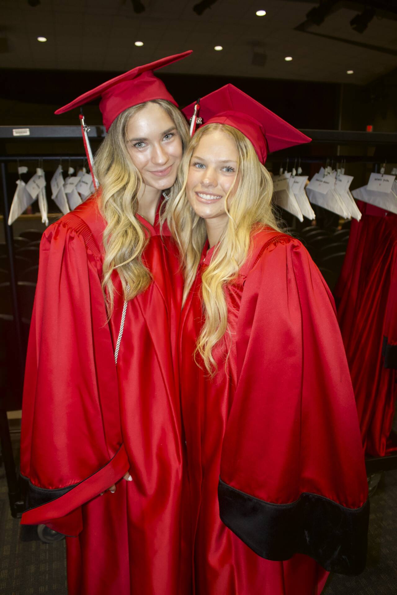 Two graduates in red caps and gowns pose together backstage before commencement.