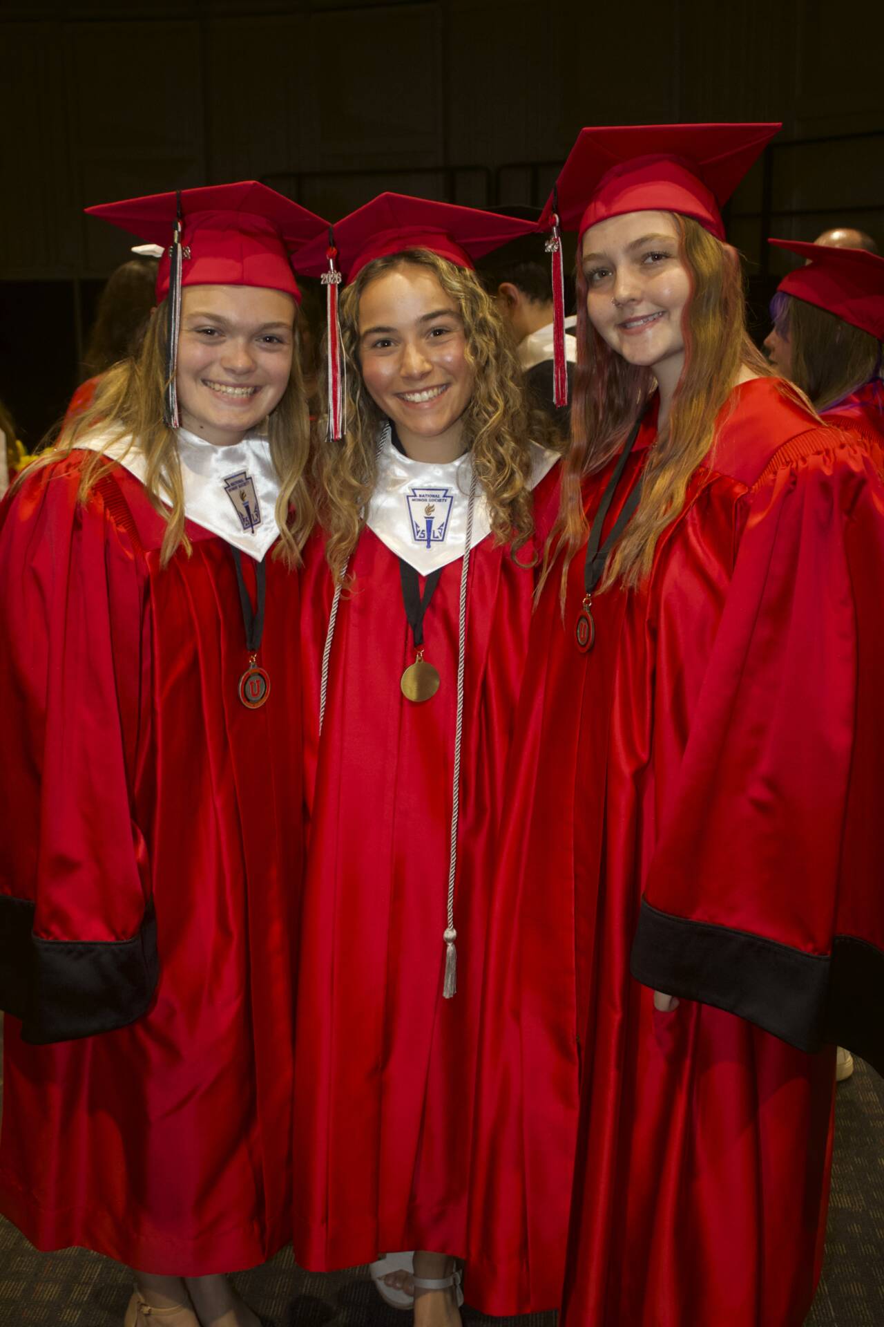 Three graduates in red caps and gowns smile together before the graduation ceremony.