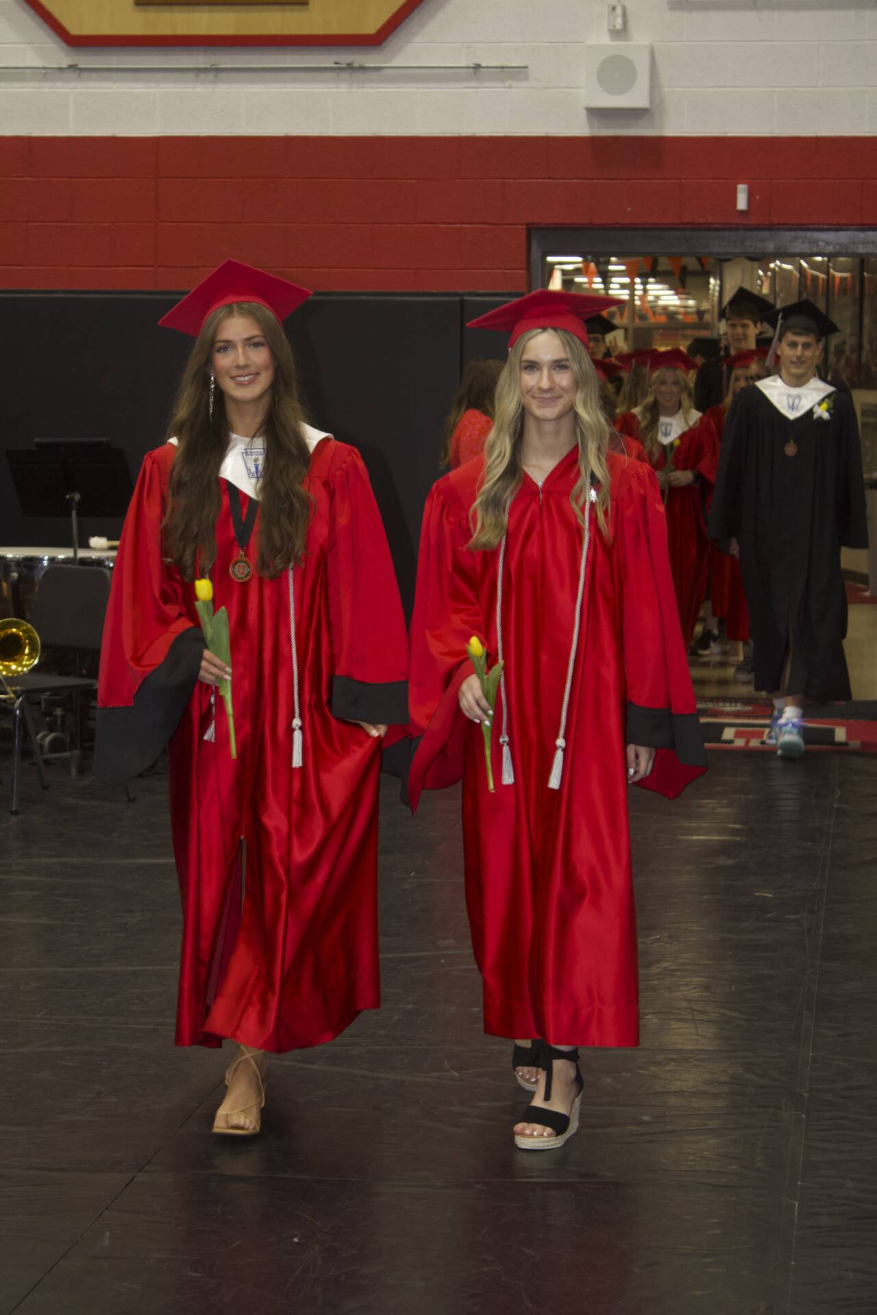 Two graduates in red caps and gowns carry yellow tulips while walking into the graduation ceremony.