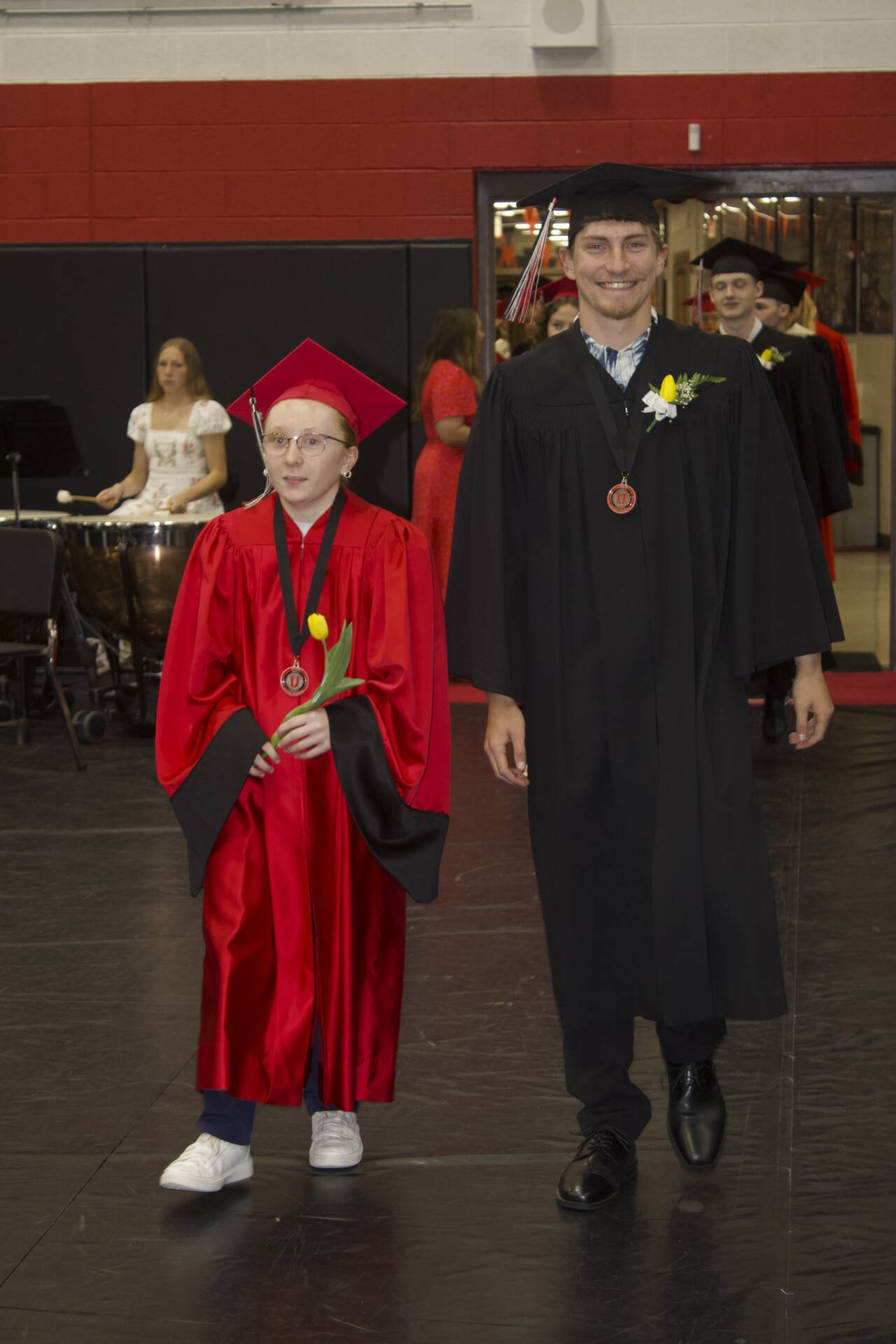Graduate in a red cap and gown walks beside a graduate in black during the commencement procession.