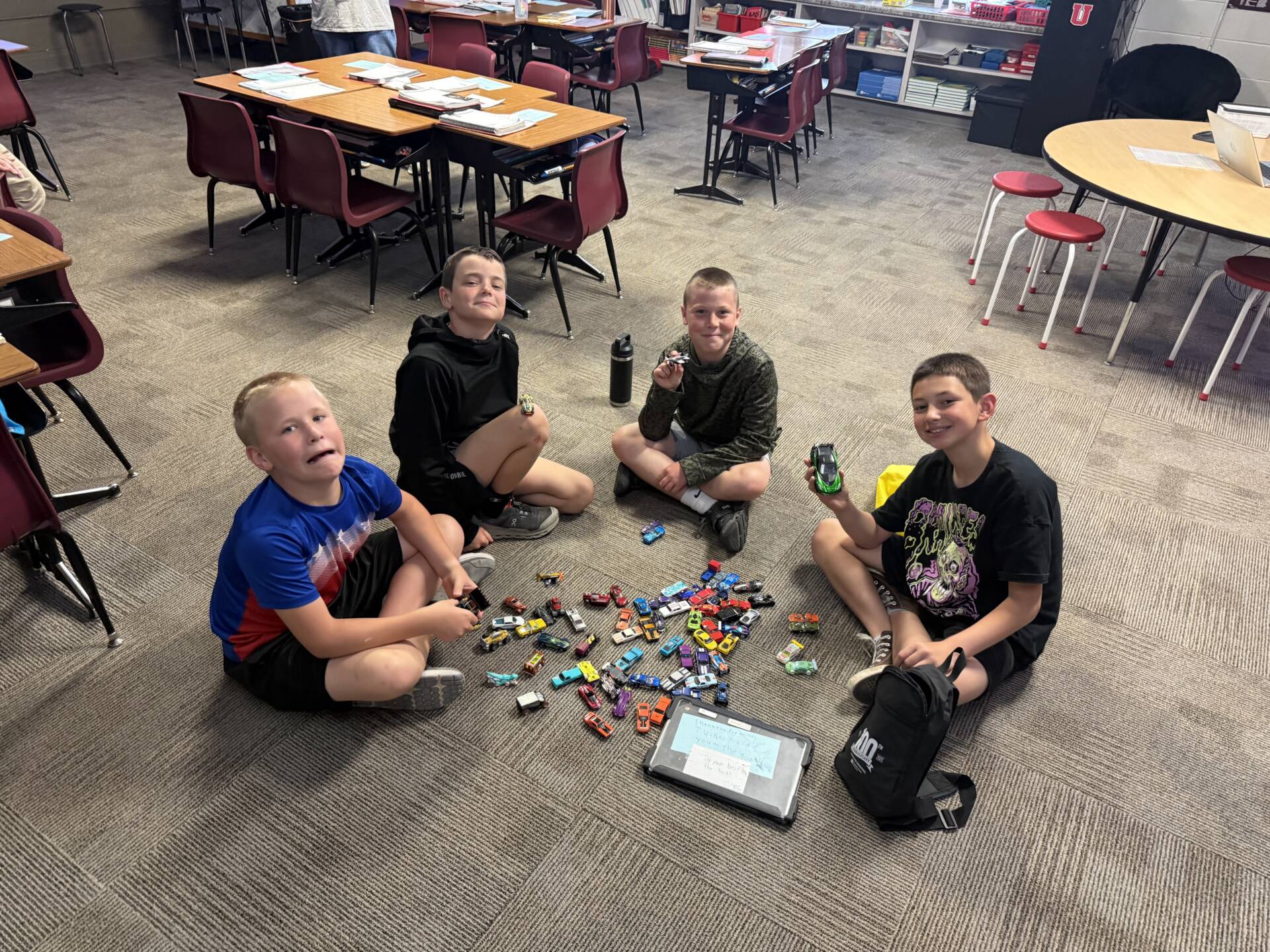 Four students sit in a circle on a classroom floor, sorting and playing with a collection of toy cars.