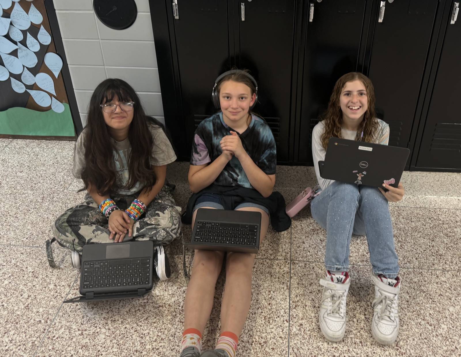 Three students sit on the hallway floor using laptops and headphones while smiling toward the camera.