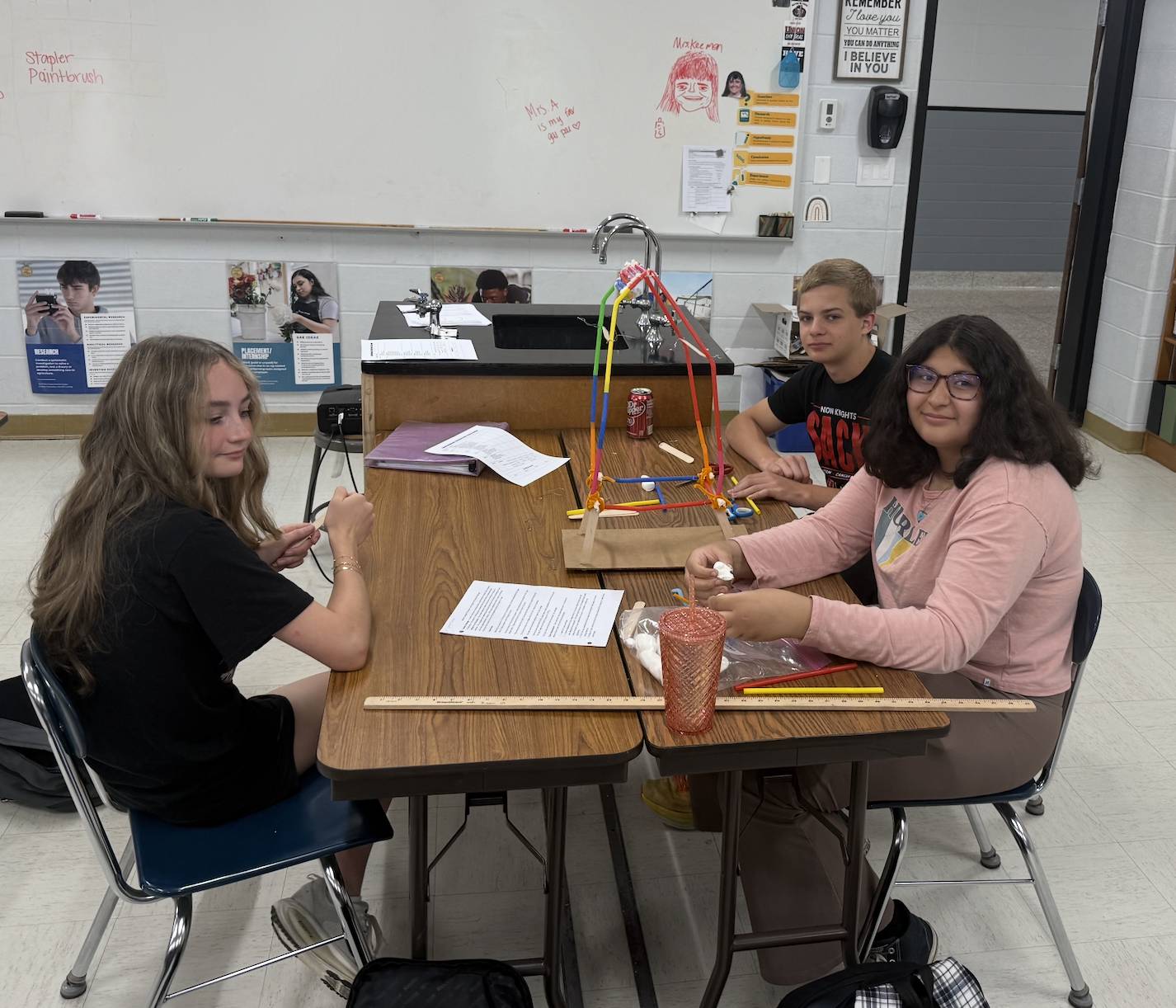 Three students build a colorful straw-and-stick tower during a classroom STEM activity at a shared table.