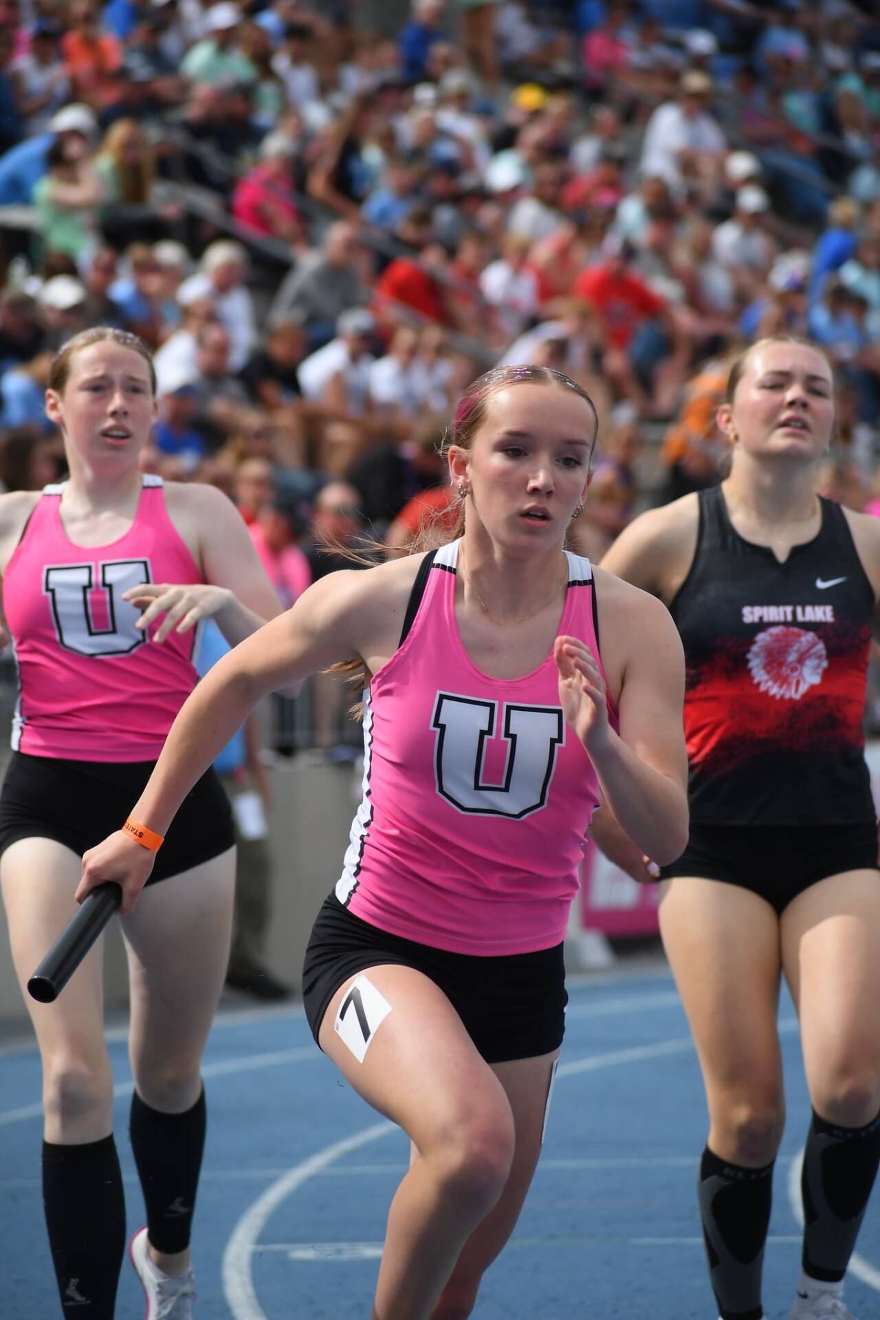 Relay runner in a pink uniform carries a baton around the curve during a track race, with competitors close behind.