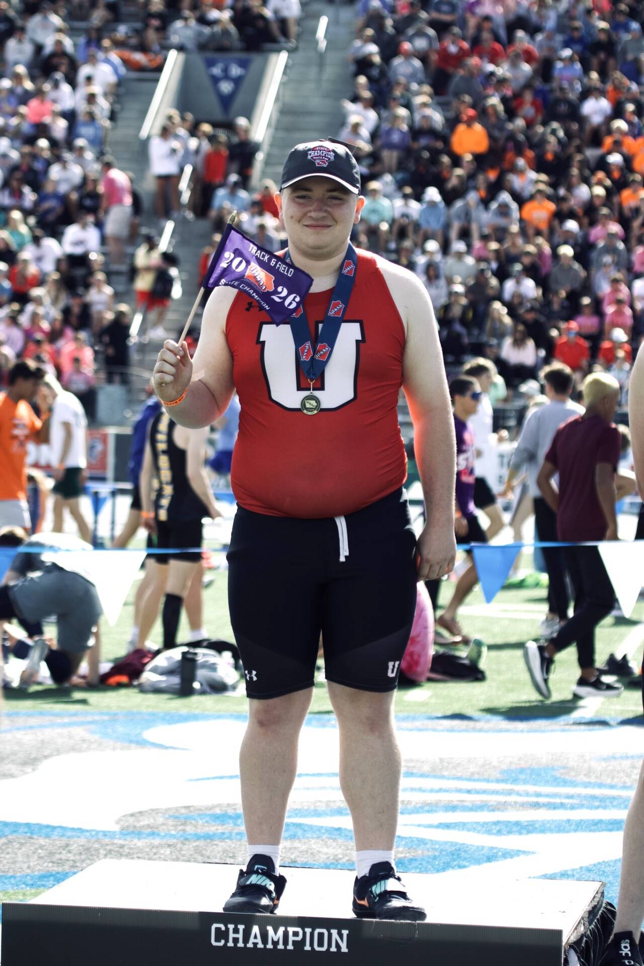 Student athlete wearing a medal stands on a champion podium, holding a state track and field champion flag in front of a crowded stadium.
