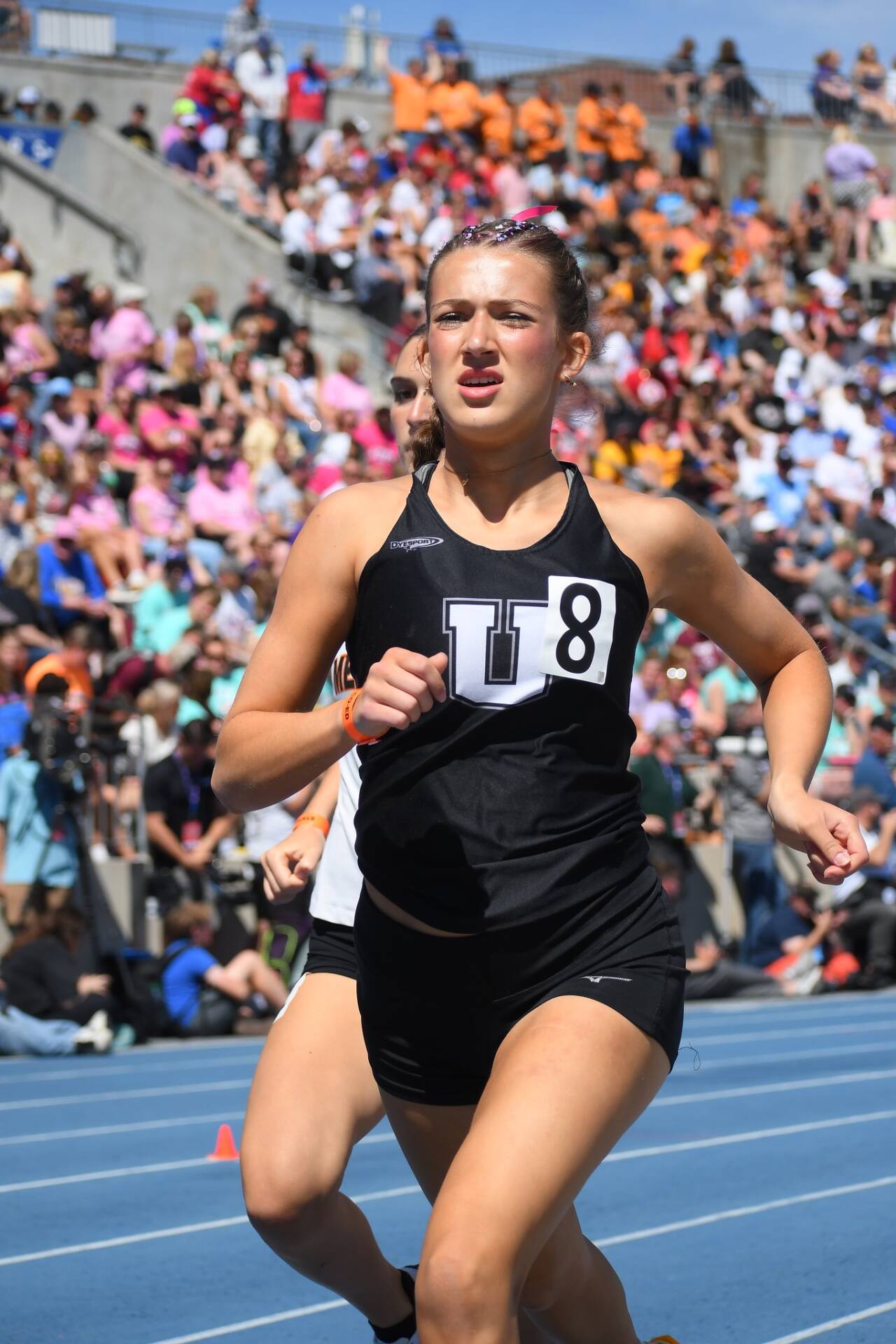 Student athlete in a black uniform races on a blue track during a state meet, with a packed stadium of spectators in the background.