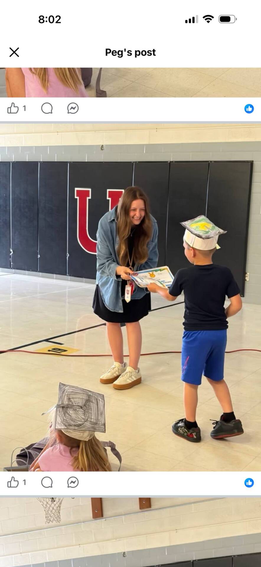 Teacher presents a certificate to a smiling student wearing a handmade graduation cap during an elementary school promotion ceremony.