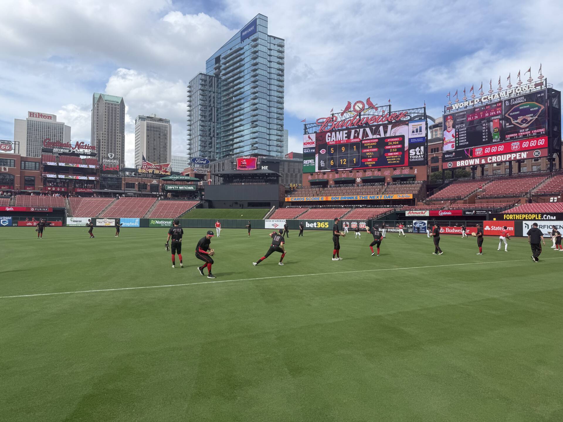 Players warm up on the field at a major league baseball stadium, with the city skyline and scoreboard visible in the background.