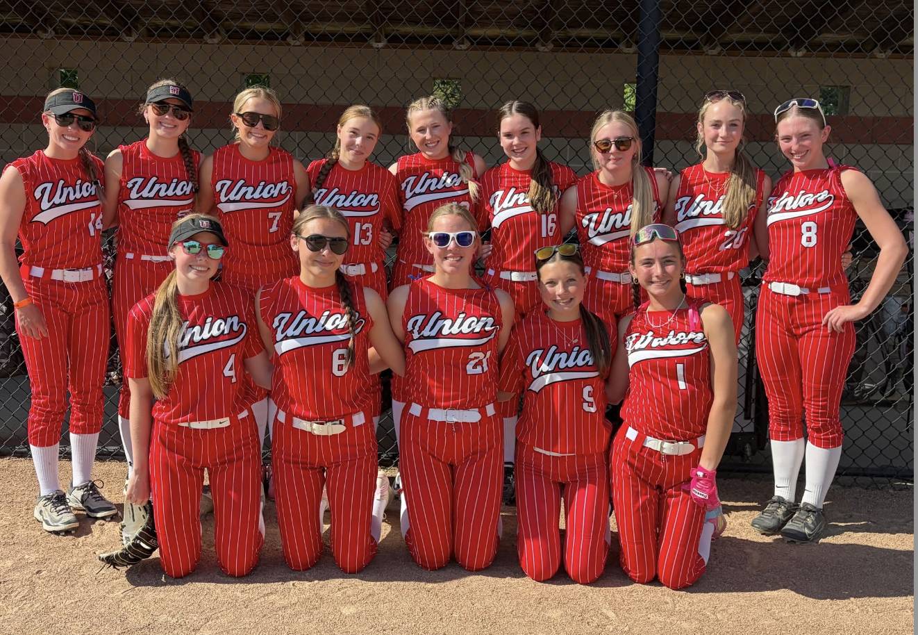 Middle school softball players in red Union uniforms pose together in front of a dugout on a sunny day.