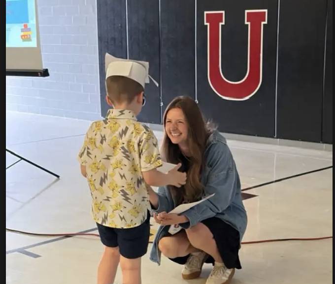 A young student in a paper graduation cap receives a certificate while smiling with a kneeling teacher during a school ceremony.
