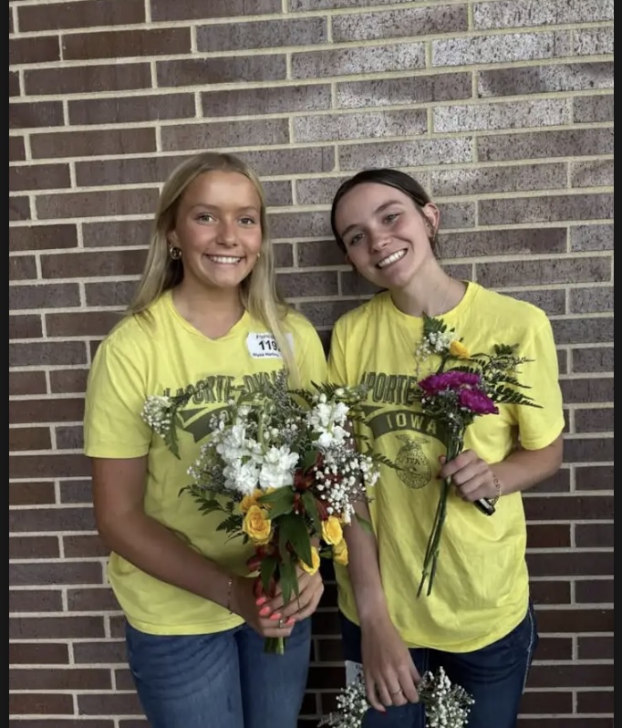 Two FFA members smile and hold floral arrangements while posing in front of a brick wall.