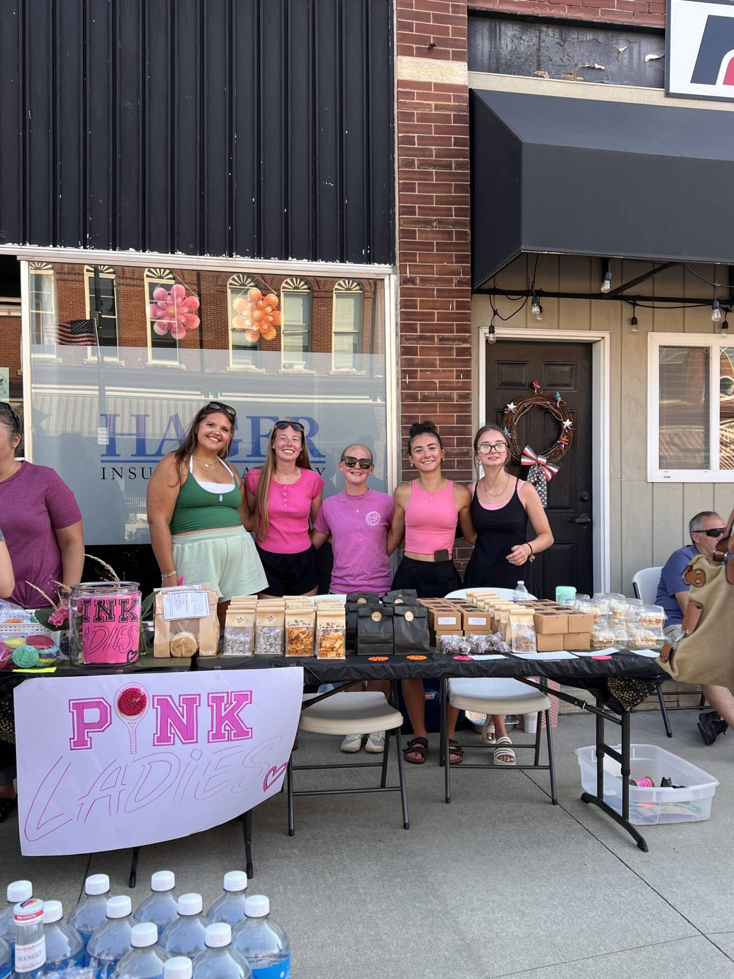 Five young women stand behind a table of baked goods beneath a handmade “Pink Ladies” sign at an outdoor event.
