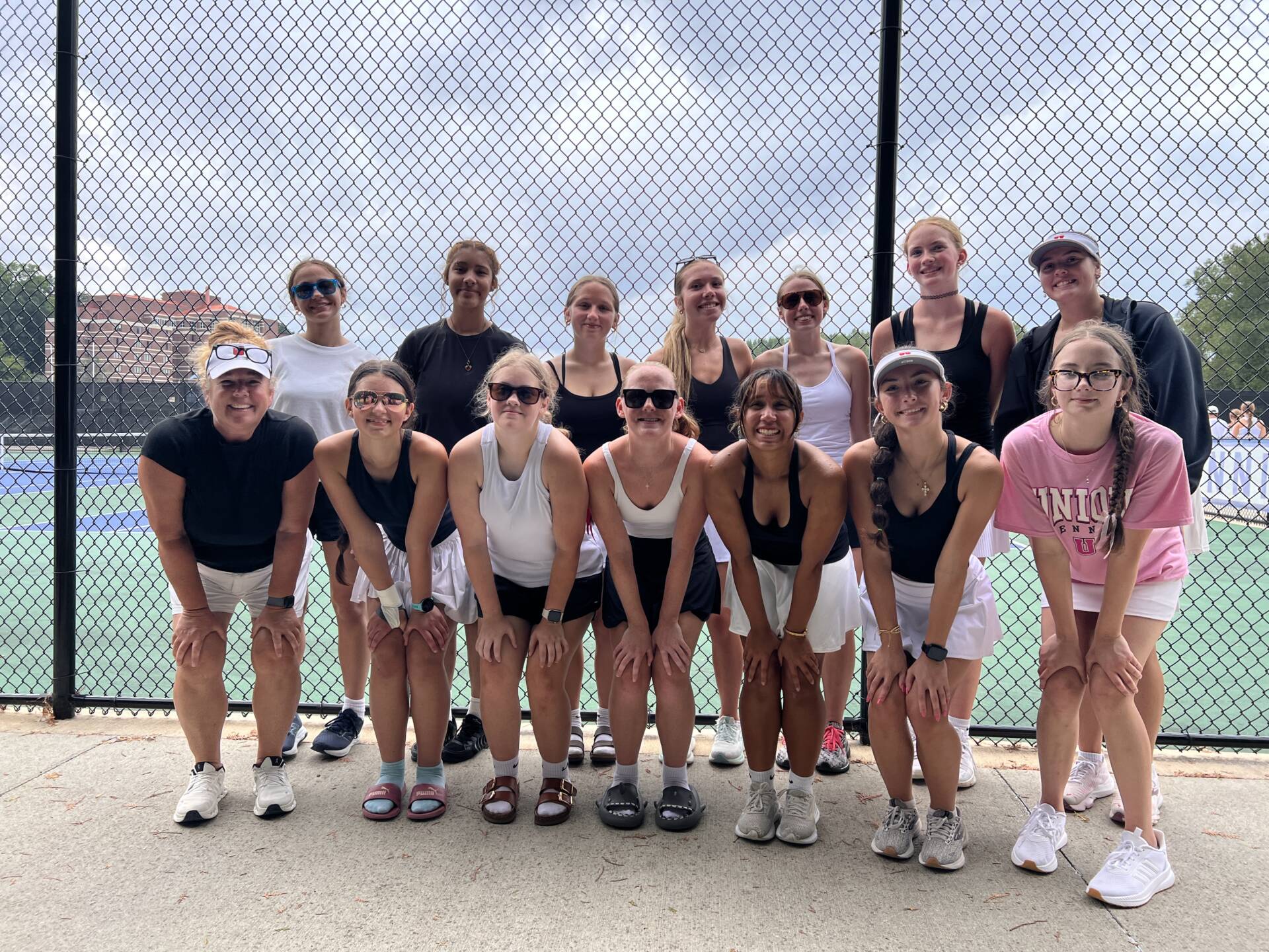 Group of tennis players and a coach pose together beside outdoor courts after a match or practice.