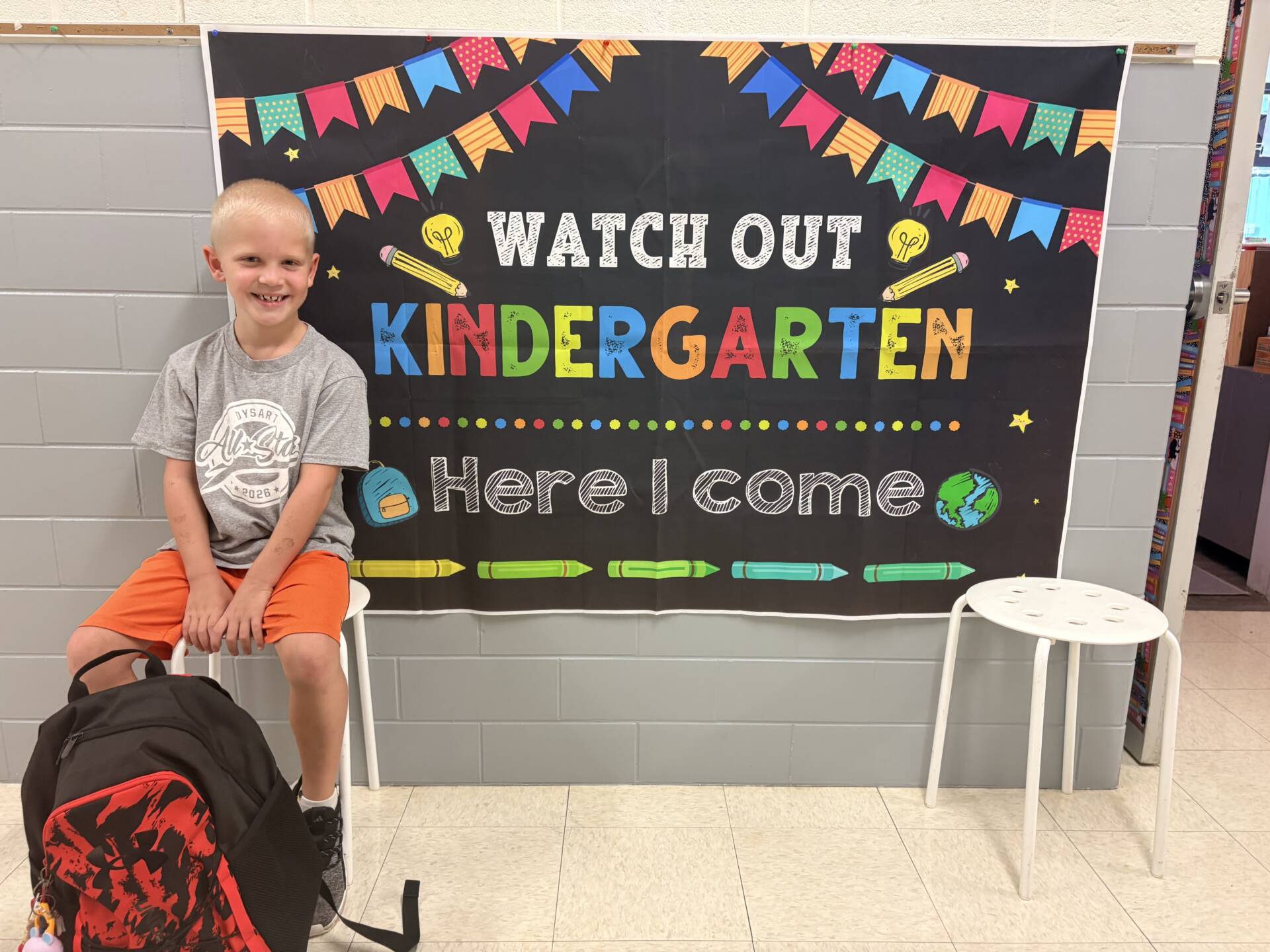 Smiling kindergartner sits beside a colorful “Watch Out Kindergarten, Here I Come” bulletin board with his backpack.