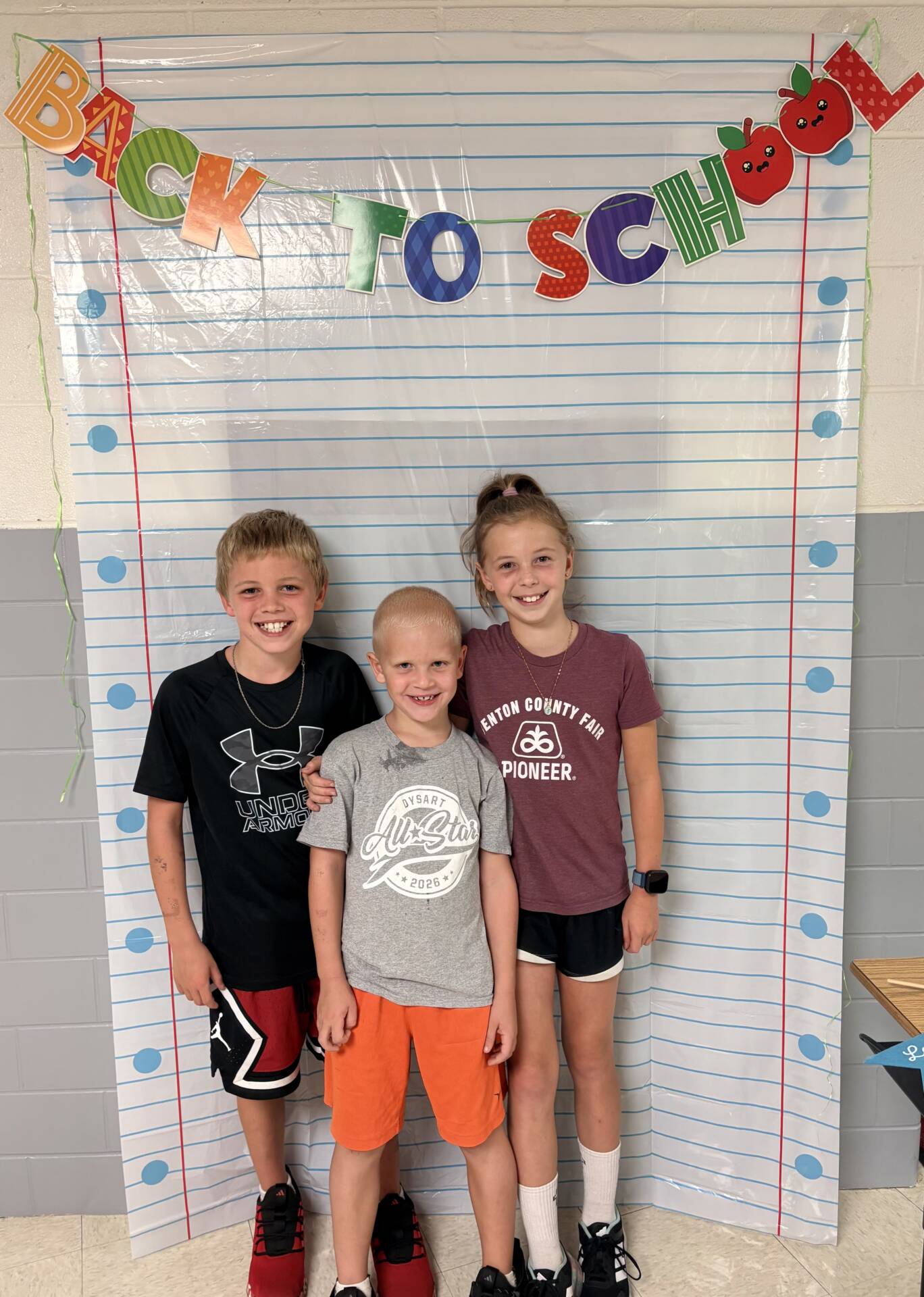 Three smiling children pose together in front of a colorful, school-themed “Back to School” photo backdrop.