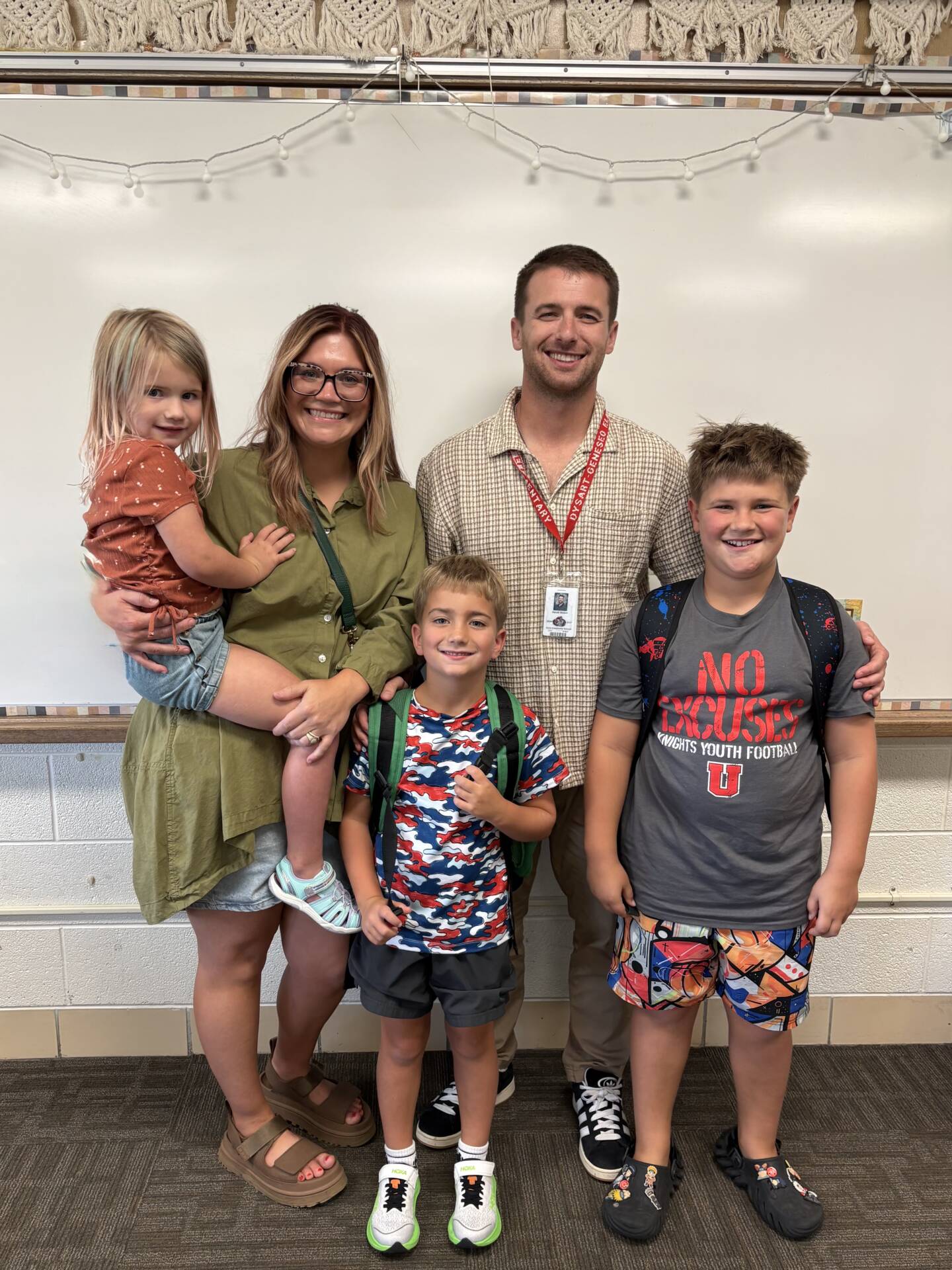 Smiling group of two adults and three children pose together in a classroom, with two children wearing backpacks.