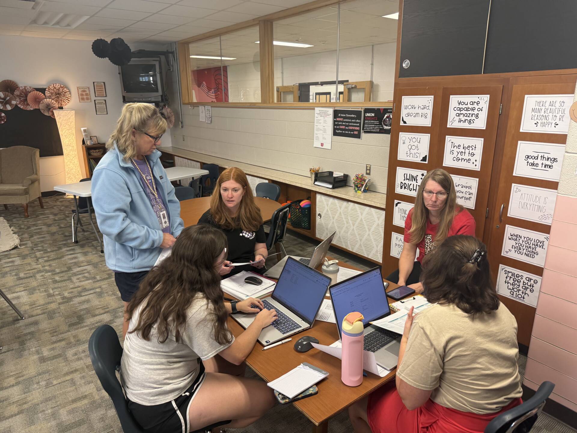Five school staff members gather around a classroom table, collaborating with laptops, notes, and printed materials.