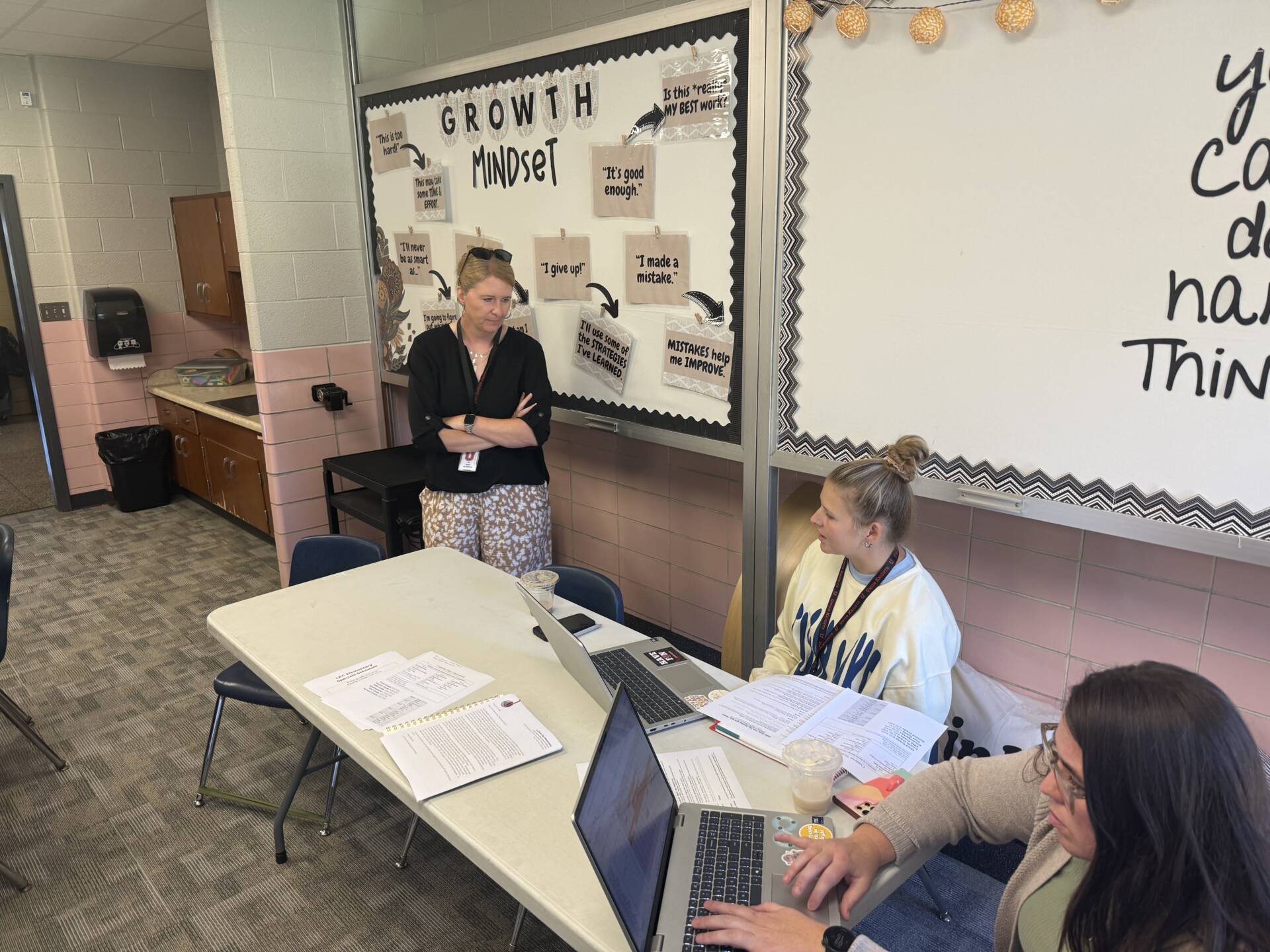 Three school staff members work with laptops and documents near a classroom bulletin board about growth mindset.