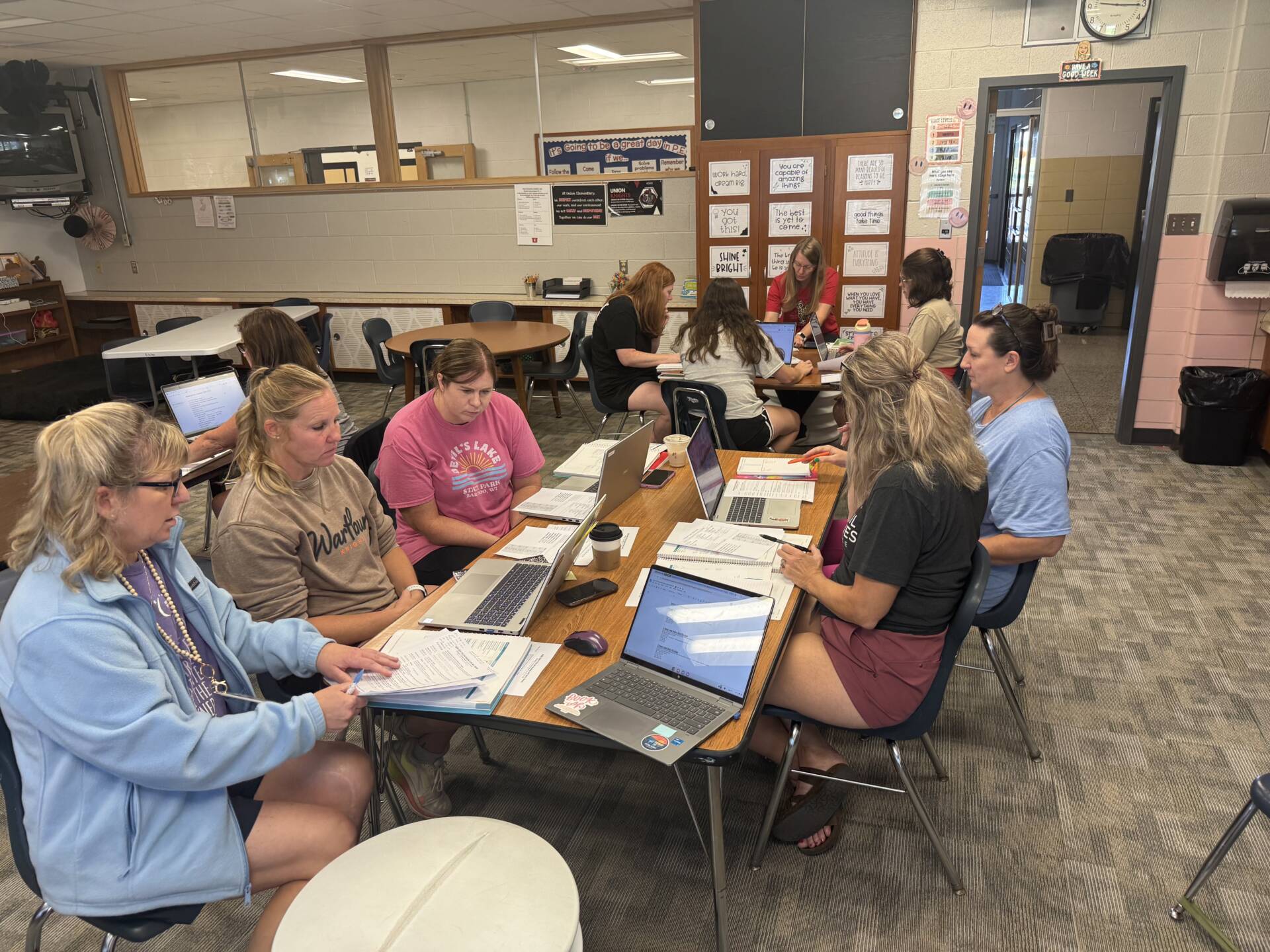 School staff collaborate around classroom tables using laptops, printed materials, notebooks, and other planning resources.