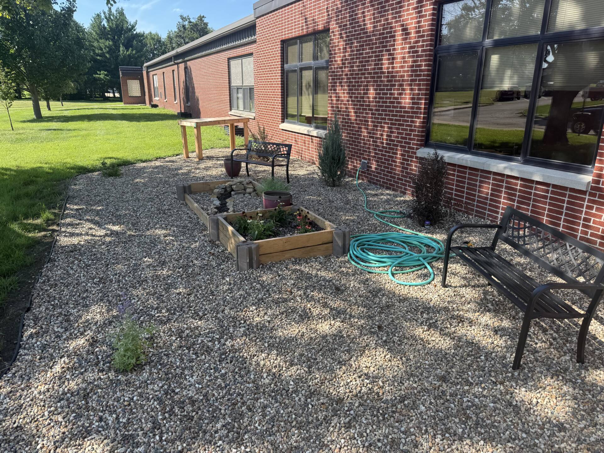Outdoor school garden beside a brick building, with raised flower beds, gravel, benches, and a coiled garden hose.