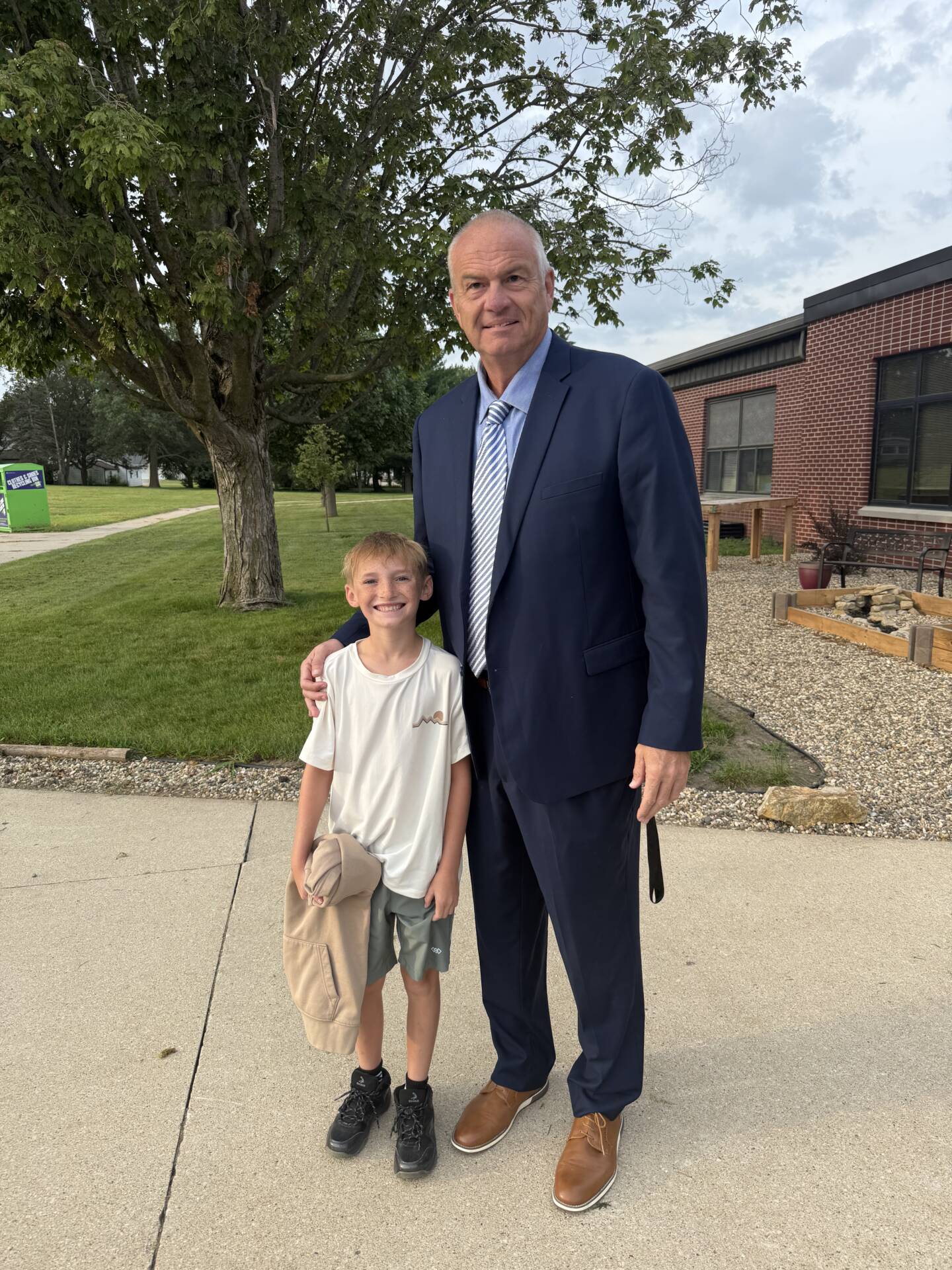 A smiling student poses outdoors beside a man in a navy suit near a brick school building.