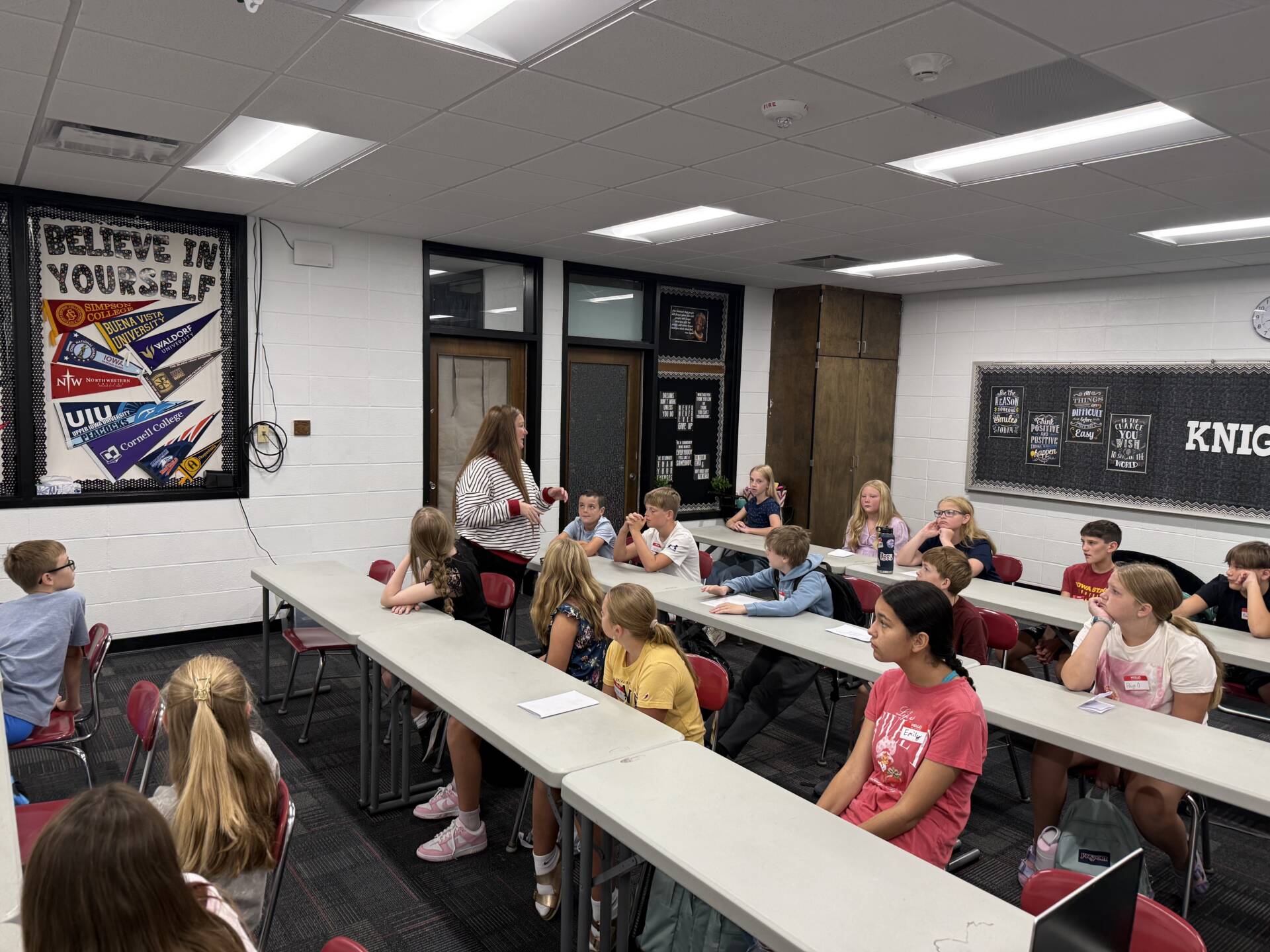 A teacher addresses students seated at classroom tables as they listen and participate in a group discussion.