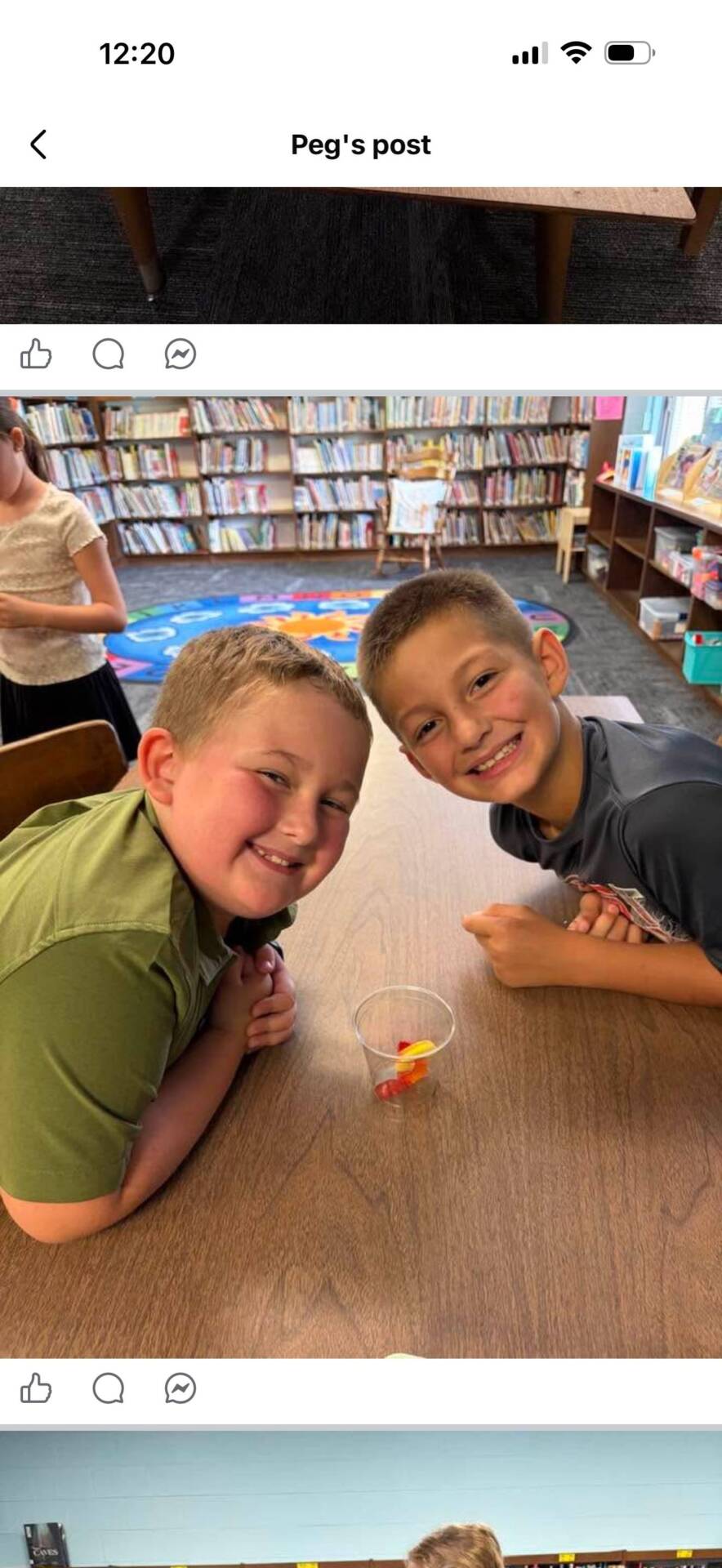 Two smiling students lean across a library table with a small cup of colorful items between them.