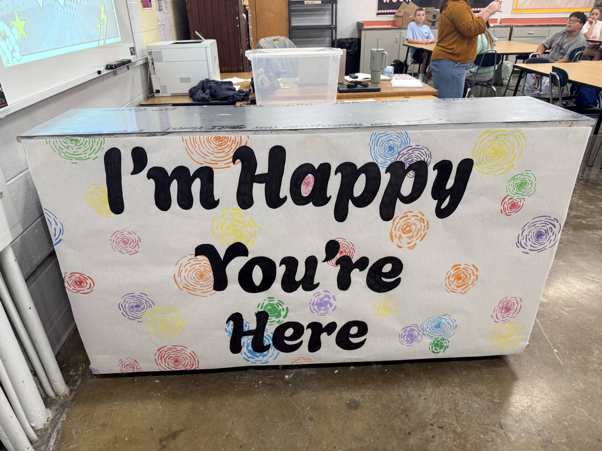 A classroom counter decorated with colorful swirls reads, “I’m Happy You’re Here,” welcoming students.